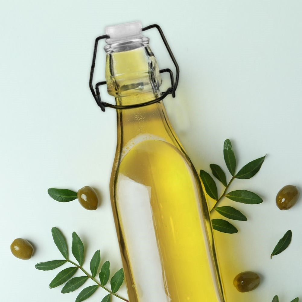 Botella de vidrio con aceite amarillo rodeada de aceitunas y hojas verdes.