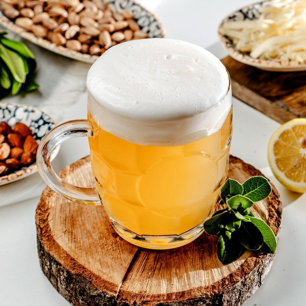 Un vaso de cerveza clara con espuma blanca sobre una tabla de madera, con frutos secos, limón y hierbas.