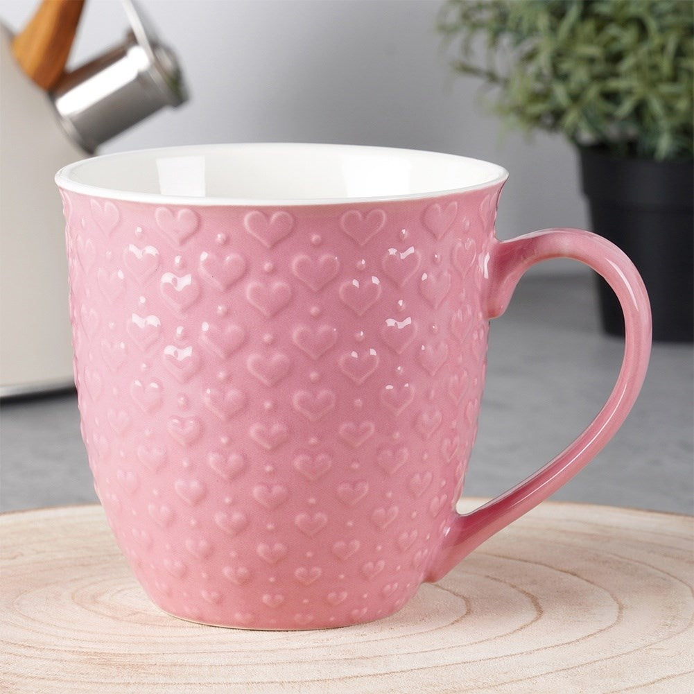 Una taza rosa con patrón de corazón sobre tabla de madera. Planta al fondo.