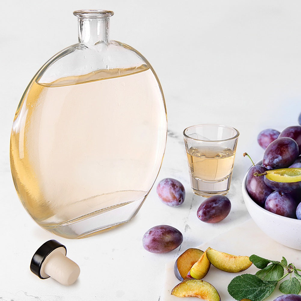 Una botella de vidrio con licor, un vaso de chupito y ciruelas.