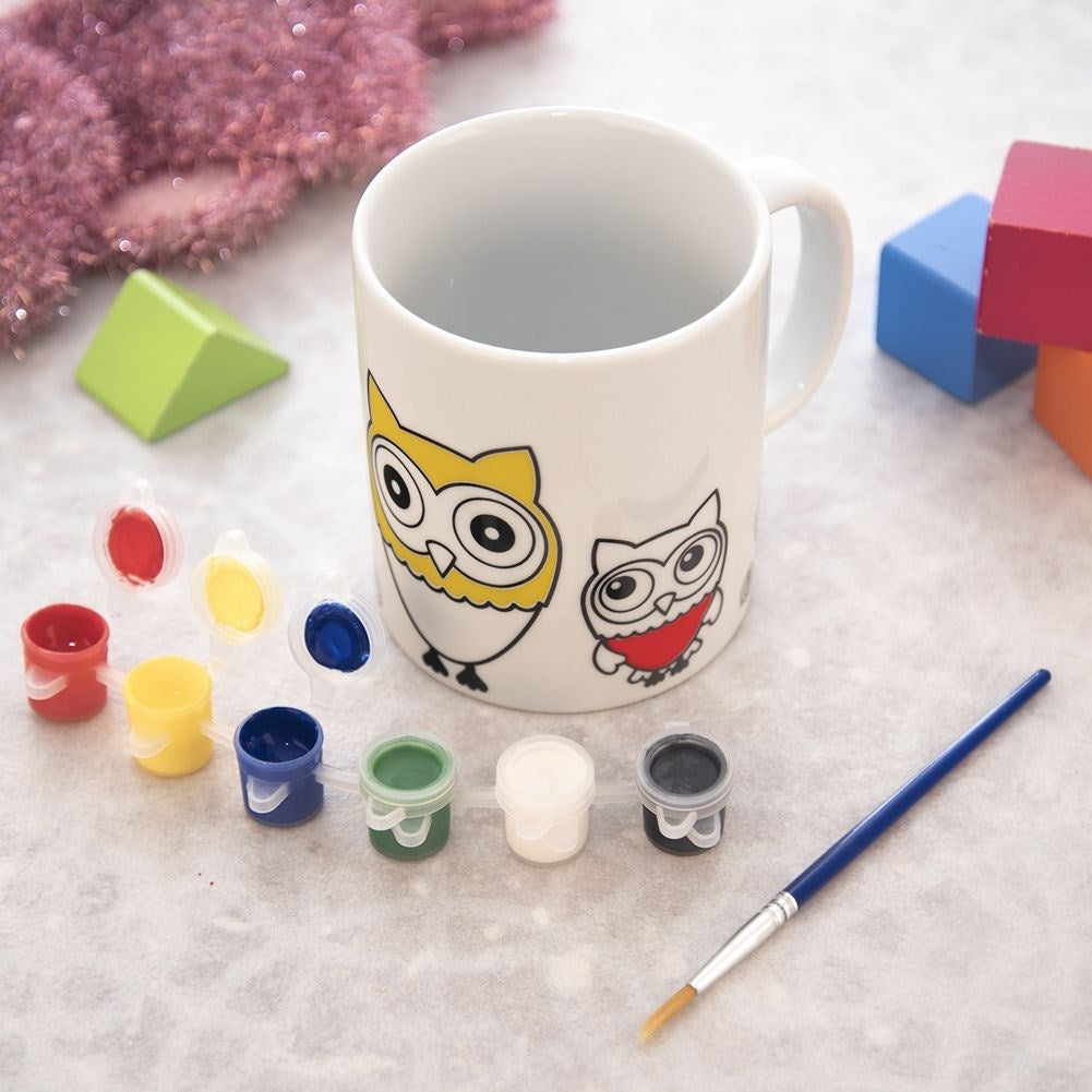 Una taza blanca con diseños de búhos, pinturas y un pincel.