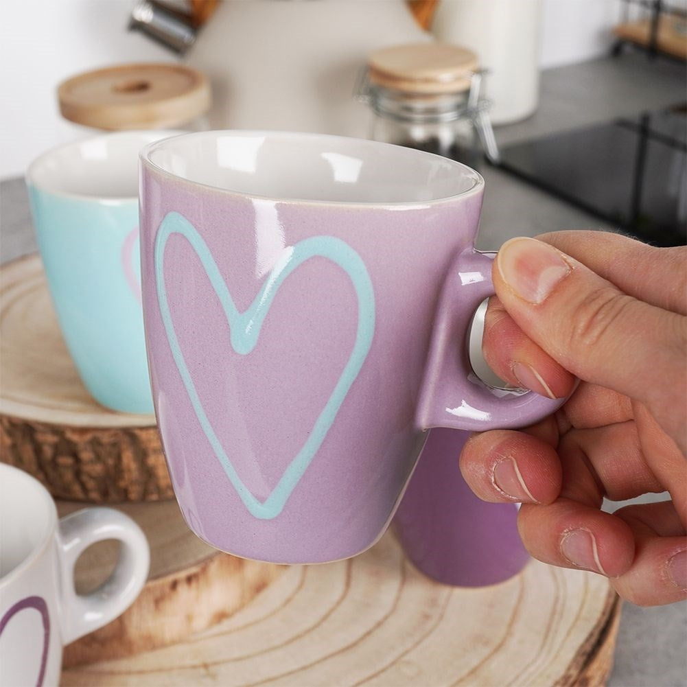 Una mano sostiene una taza morada con un corazón azul claro. Otra taza al fondo.