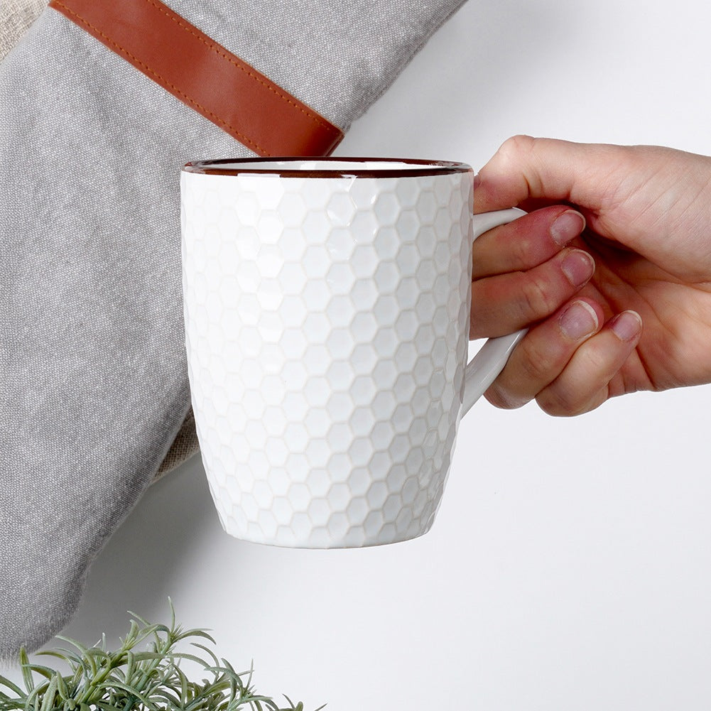 Una taza blanca con diseño de panal es sostenida por una mano, junto a un paño gris.
