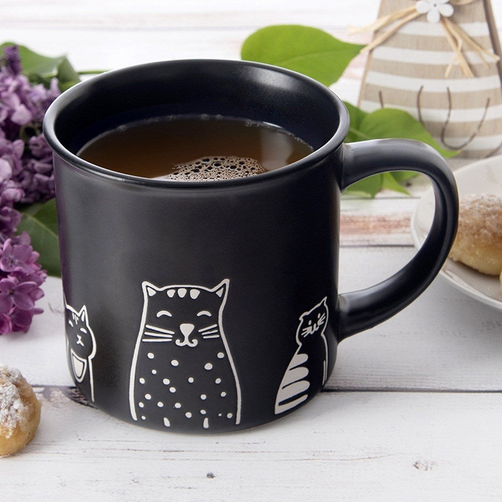 Taza negra con diseño de gato, llena de té, sobre mesa de madera.