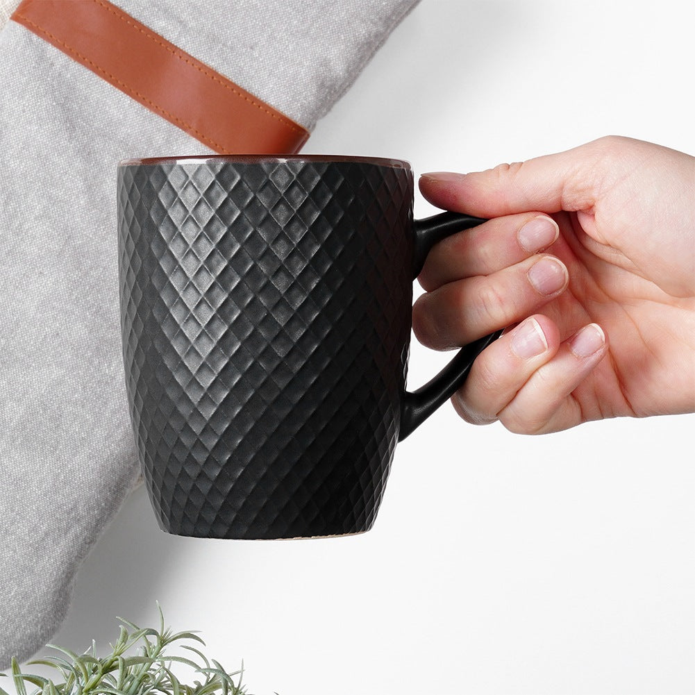 Una mano sostiene una taza negra con patrón de diamantes junto a una tela gris.