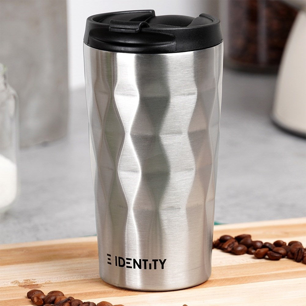 Taza de viaje plateada con tapa negra sobre superficie de madera, con granos de café cerca.