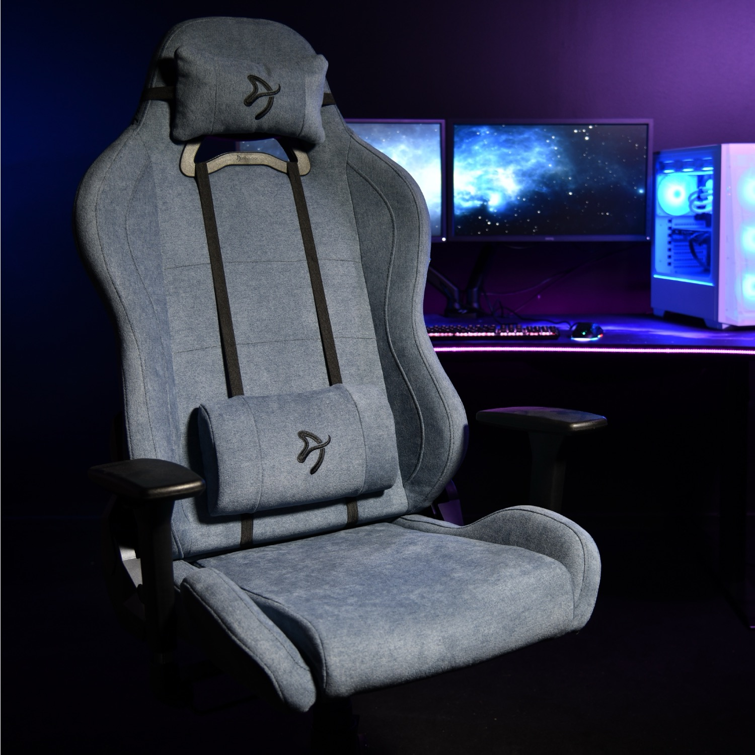 Een blauwe gamestoel met hoofd- en lendekussens staat voor een bureau met computers en monitoren.