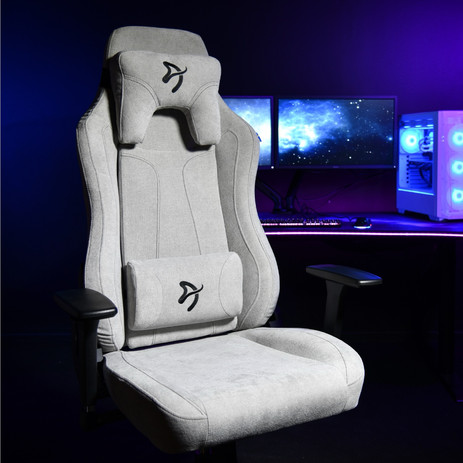 Grijze gamestoel met hoofd- en lendekussens, voor een bureau met computers.