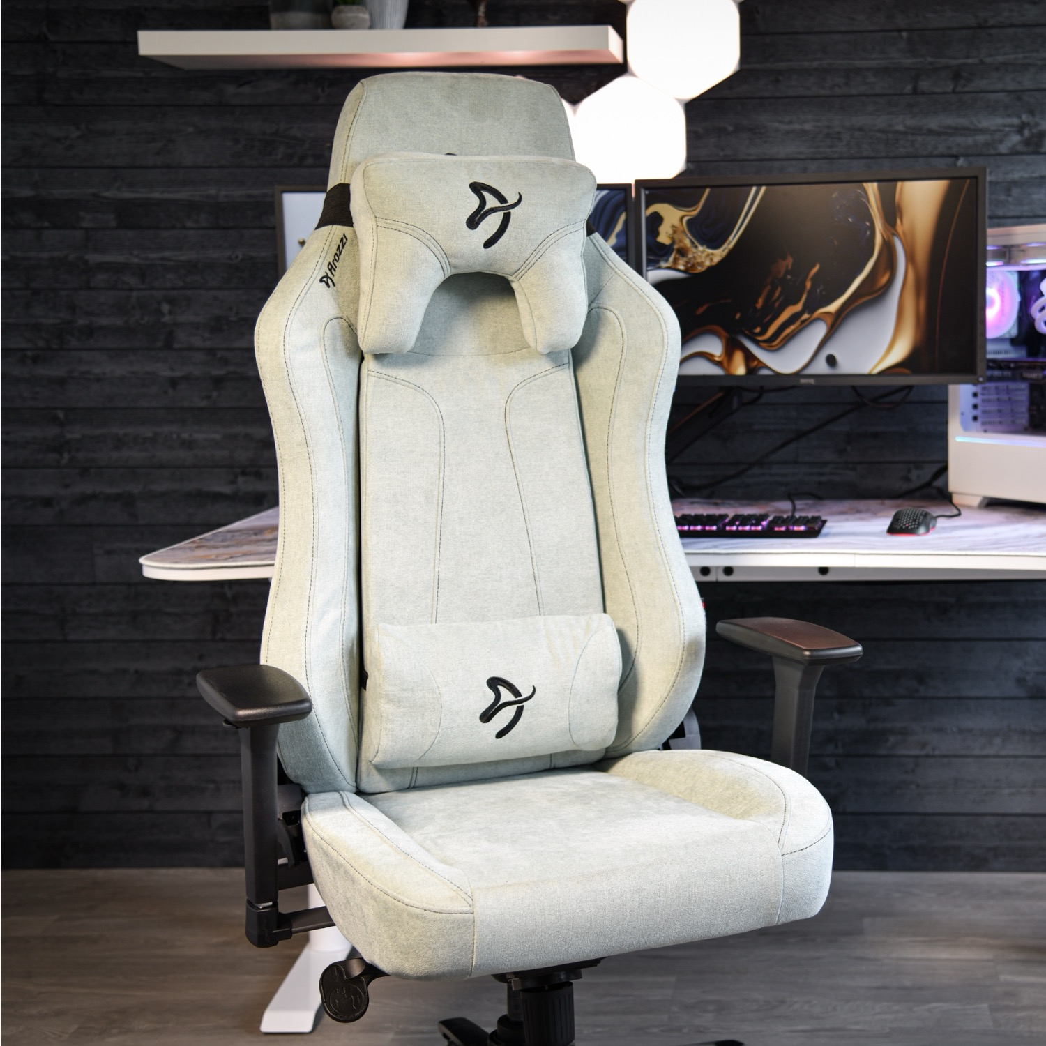 Lichtgekleurde gamestoel met nek- en lendekussens voor een bureau met monitoren.