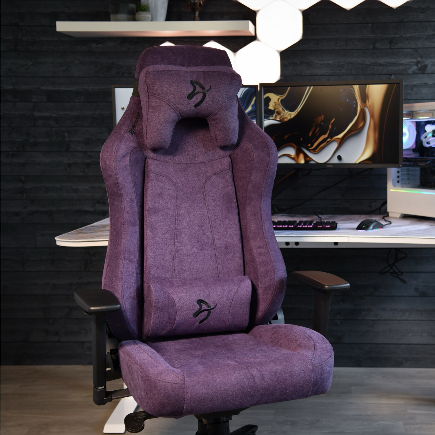 Una sedia da gaming viola con cuscini per testa e schiena di fronte a una scrivania con monitor.