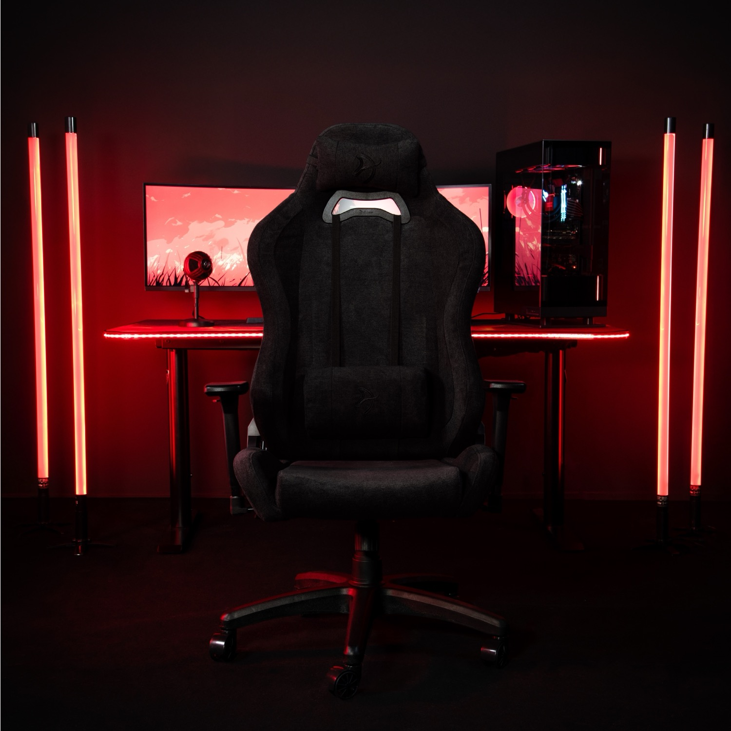 Een zwarte gamestoel staat voor een bureau met pc's en monitoren, verlicht door rode lampen.