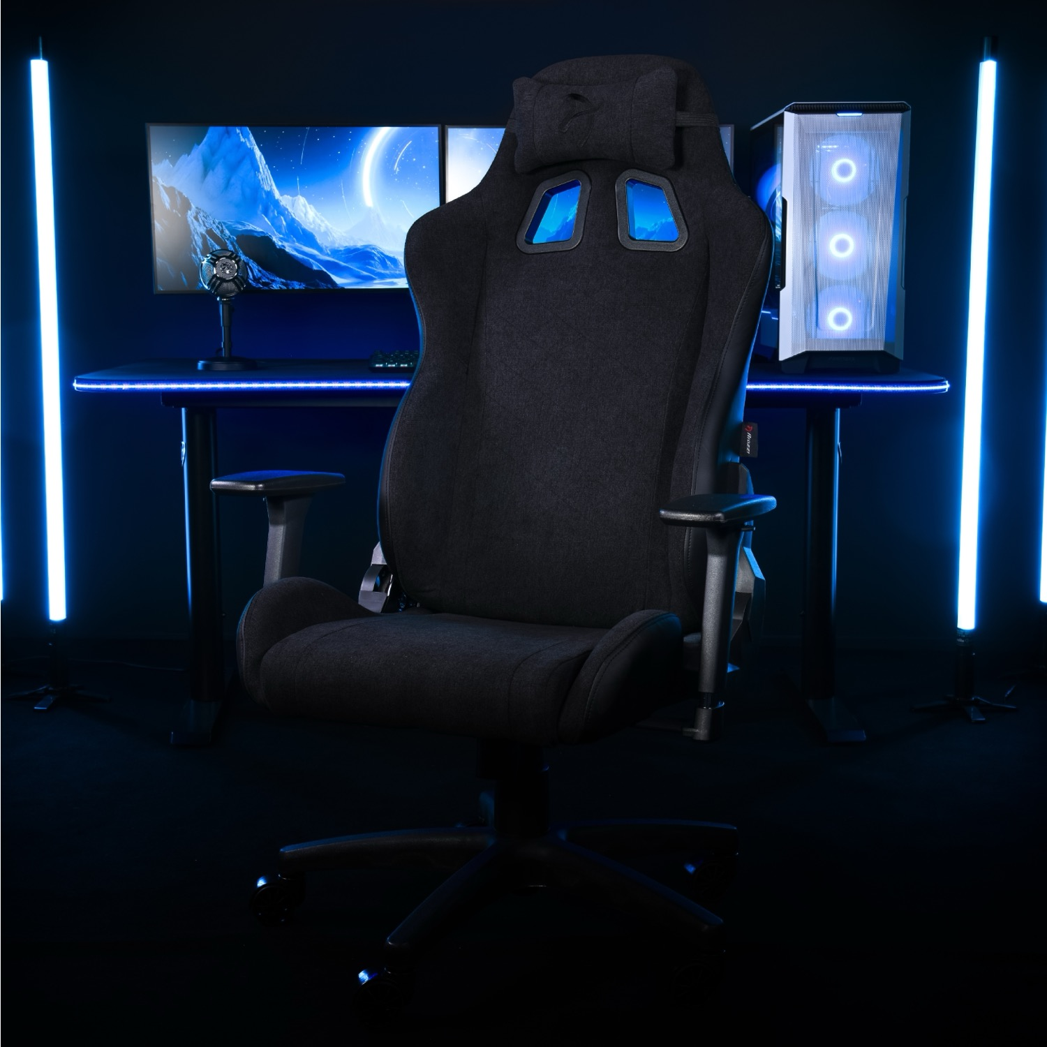 Een zwarte gamestoel voor een bureau met monitoren en pc, in blauw licht.
