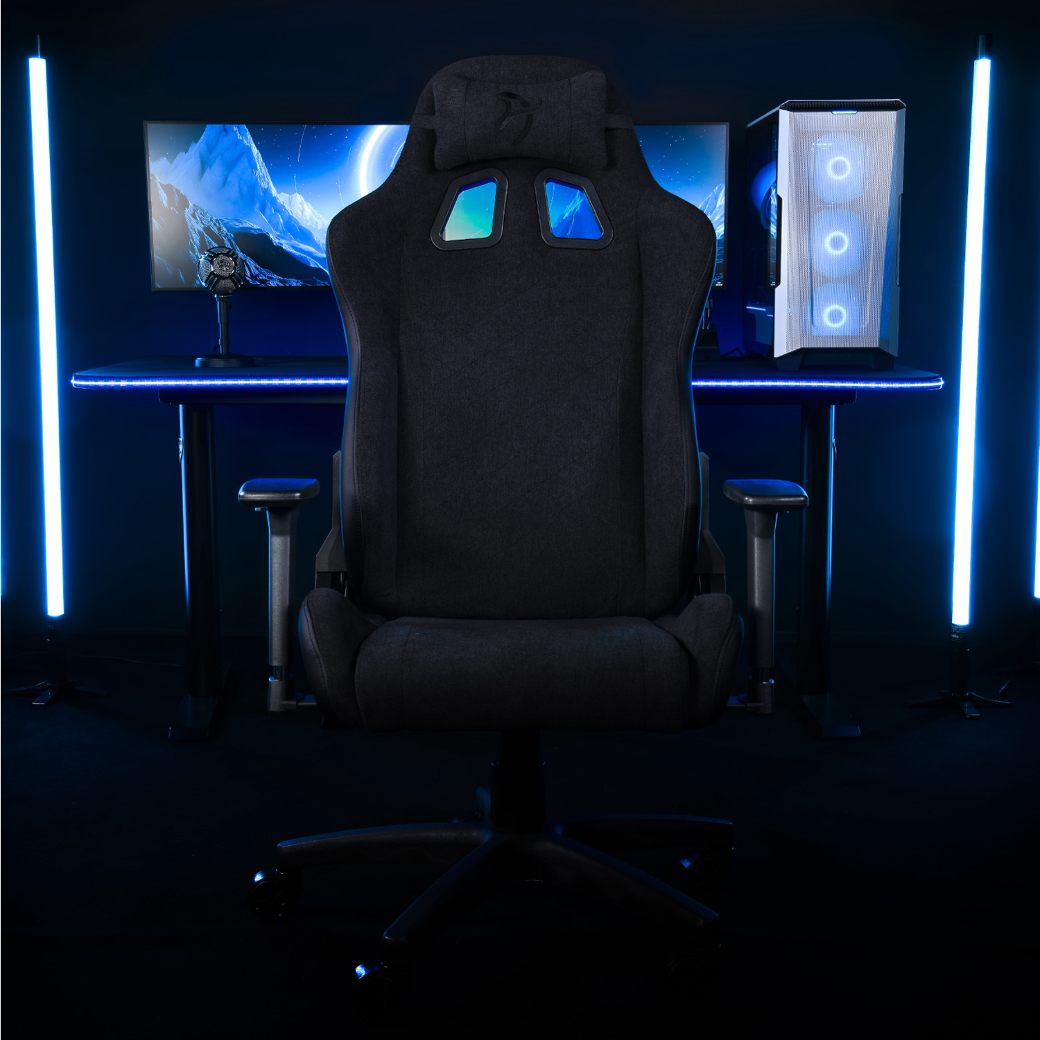 Een donkere gamestoel staat voor een bureau met monitor, computer en microfoon. Blauwe lichten flankeren de scène.