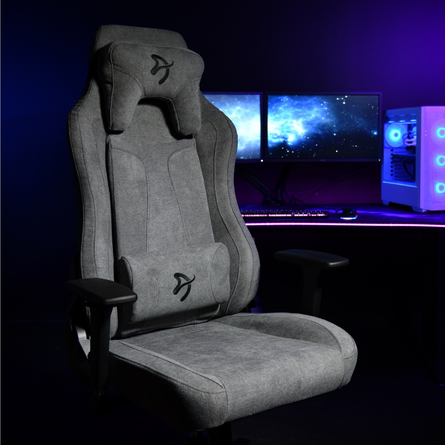 Grijze gamestoel met hoofdsteun en lendekussen voor een computer en monitoren.