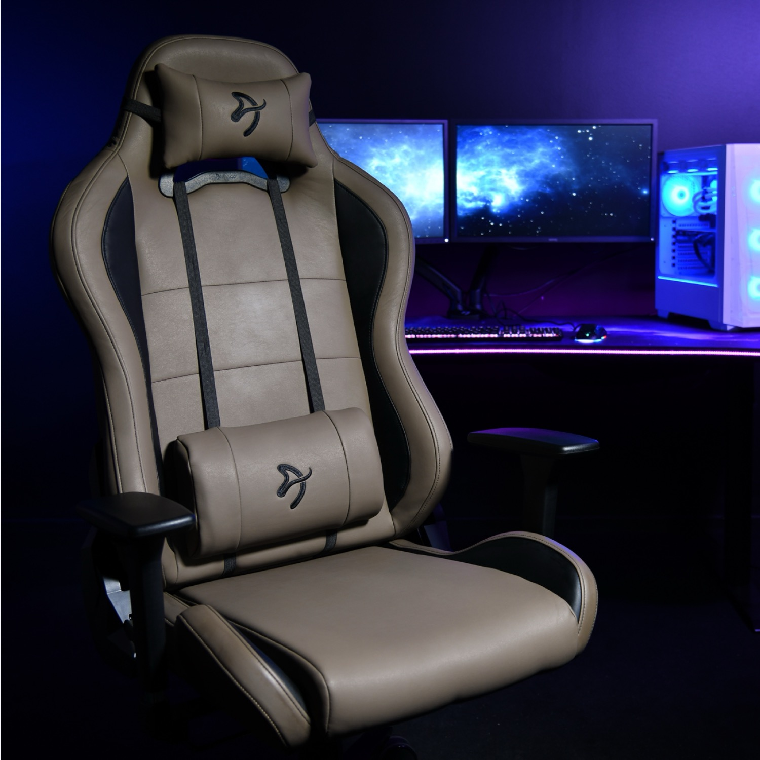 Een beige gamestoel staat voor een bureau met monitoren en een pc.