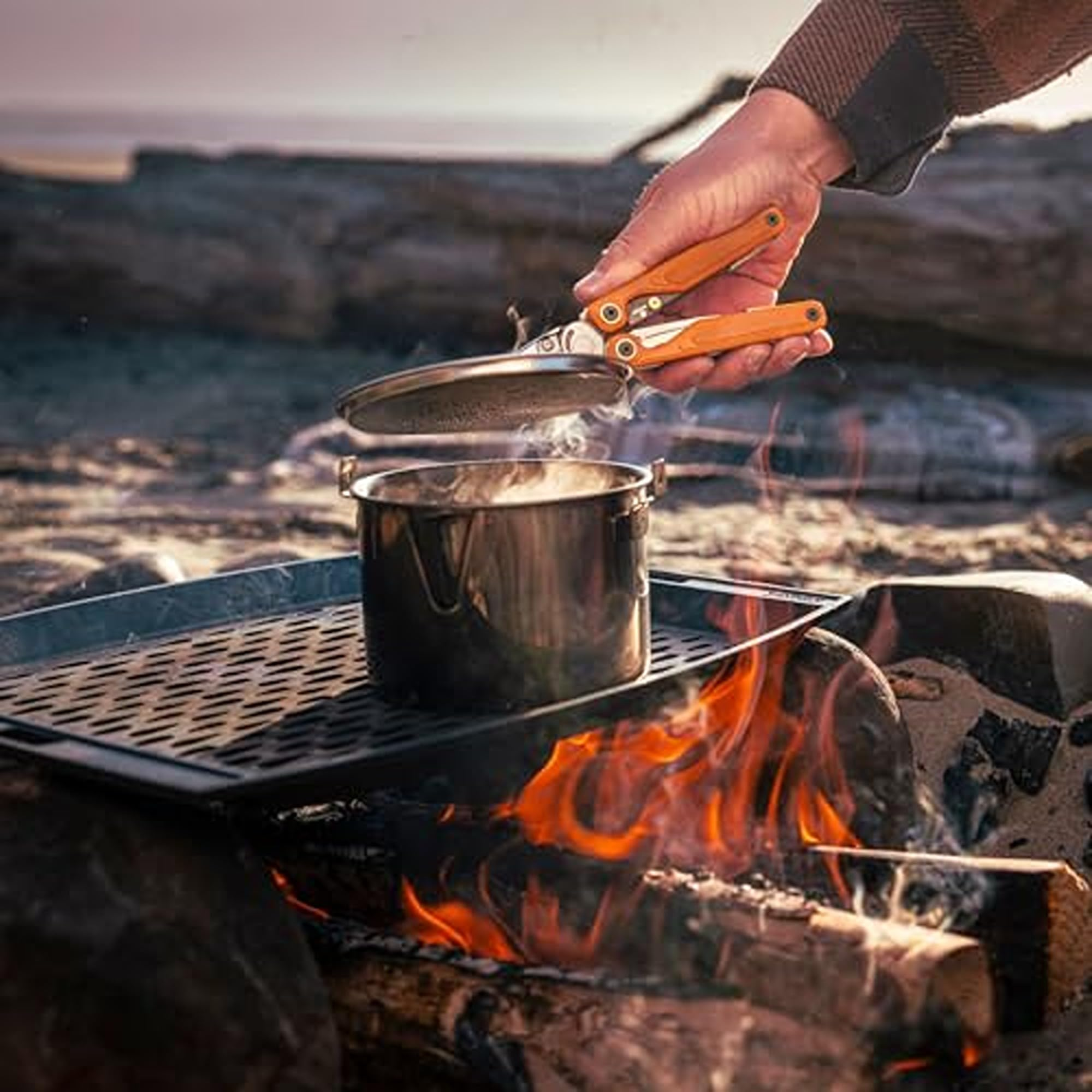 Topf wird über Feuer mit Zange gehalten, Dampf steigt auf.