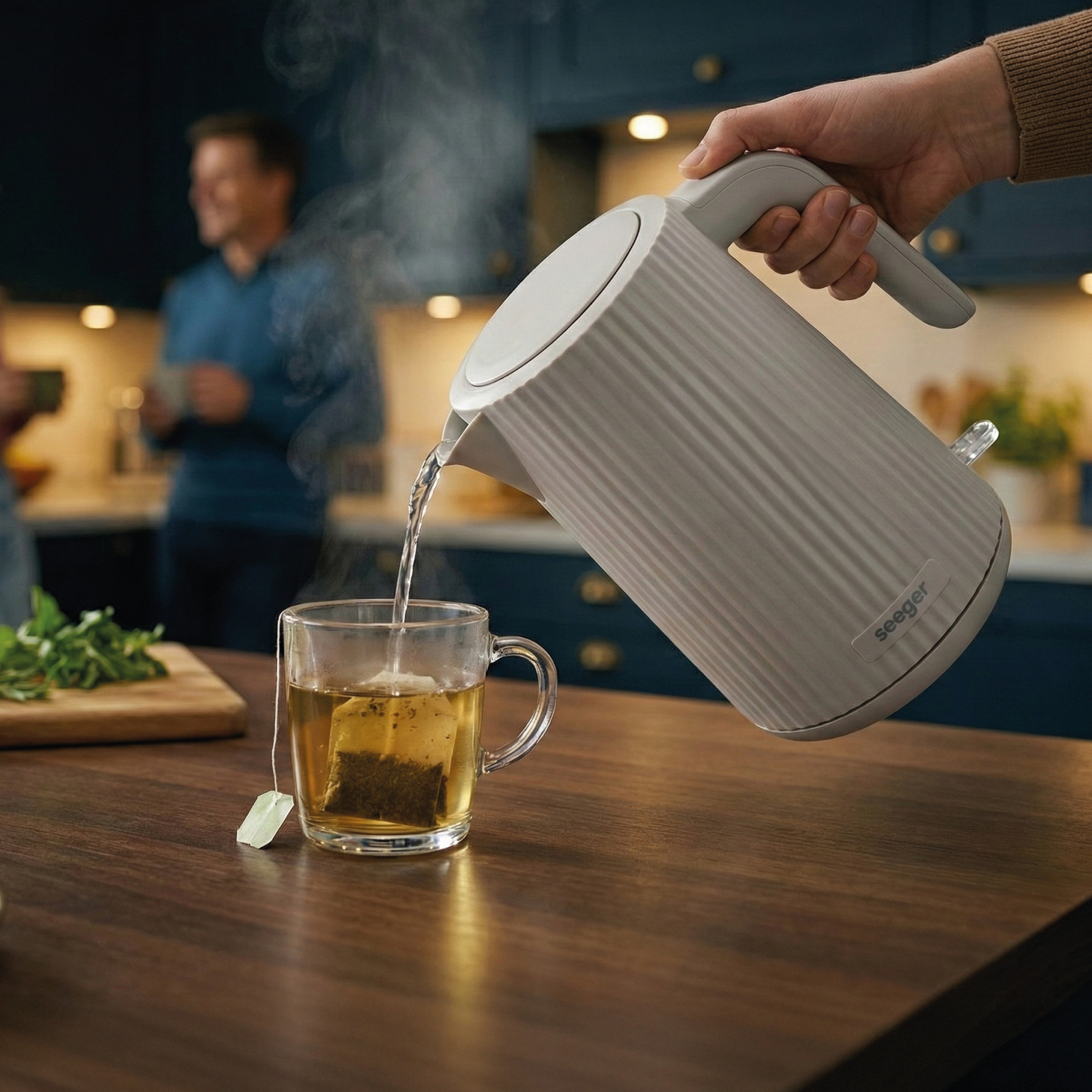 Eine Person gießt heißes Wasser aus einem grauen Wasserkocher in eine Teetasse.