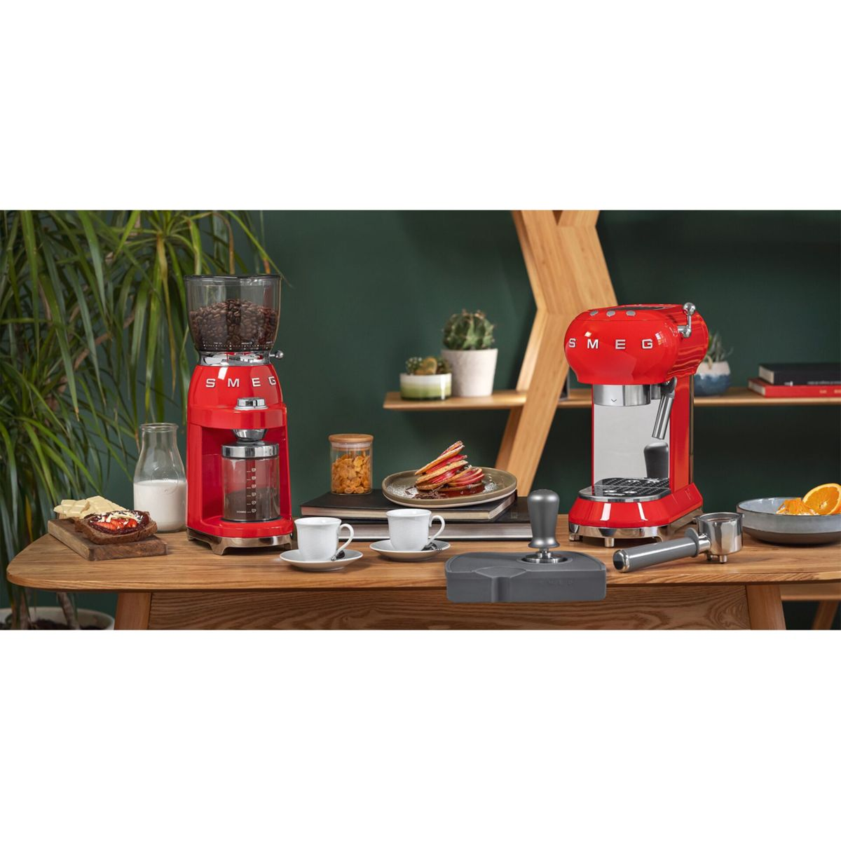 Molinillo y cafetera espresso Smeg rojos con accesorios sobre mesa de madera.