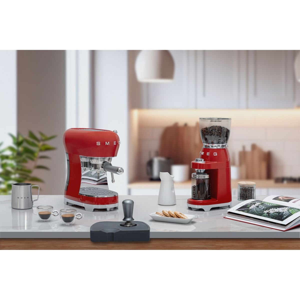Dos cafeteras Smeg rojas, una jarra de leche, tazas y galletas en una encimera.