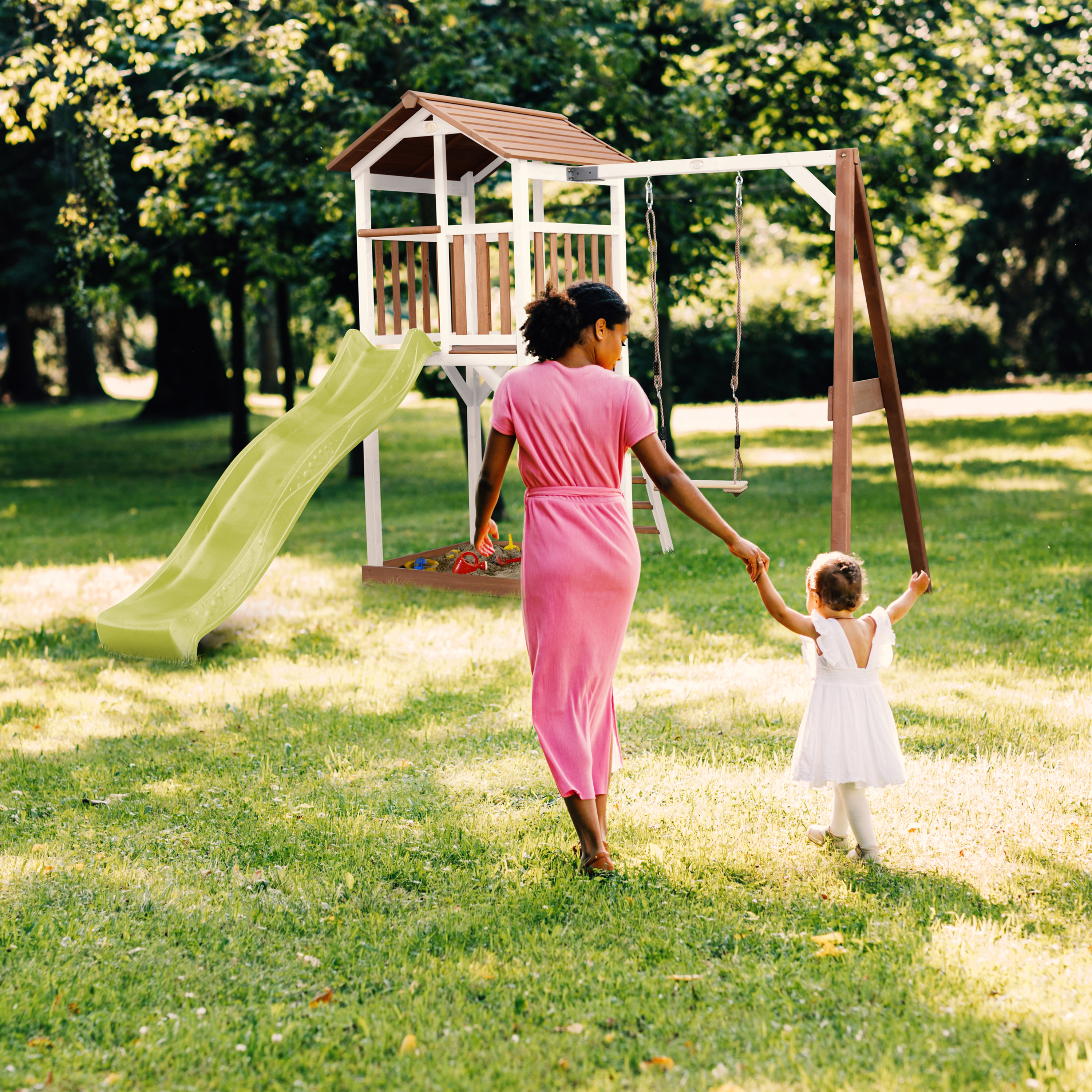 Frau in rosa Kleid und Kind in wei ßem Kleid gehen auf Spielplatz.