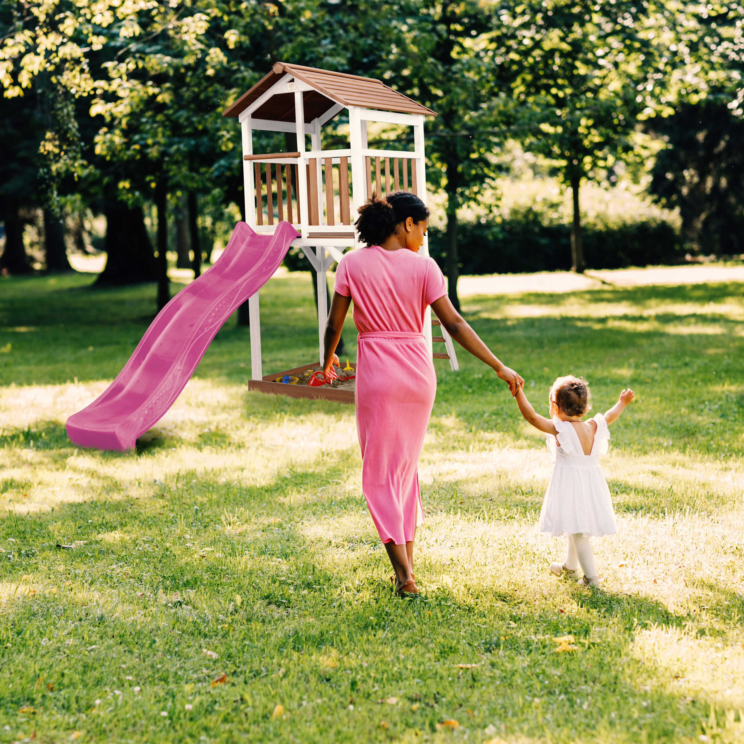 Frau und Kind gehen auf Spielplatz zu. Frau trägt rosa Kleid, Kind weißes Kleid.