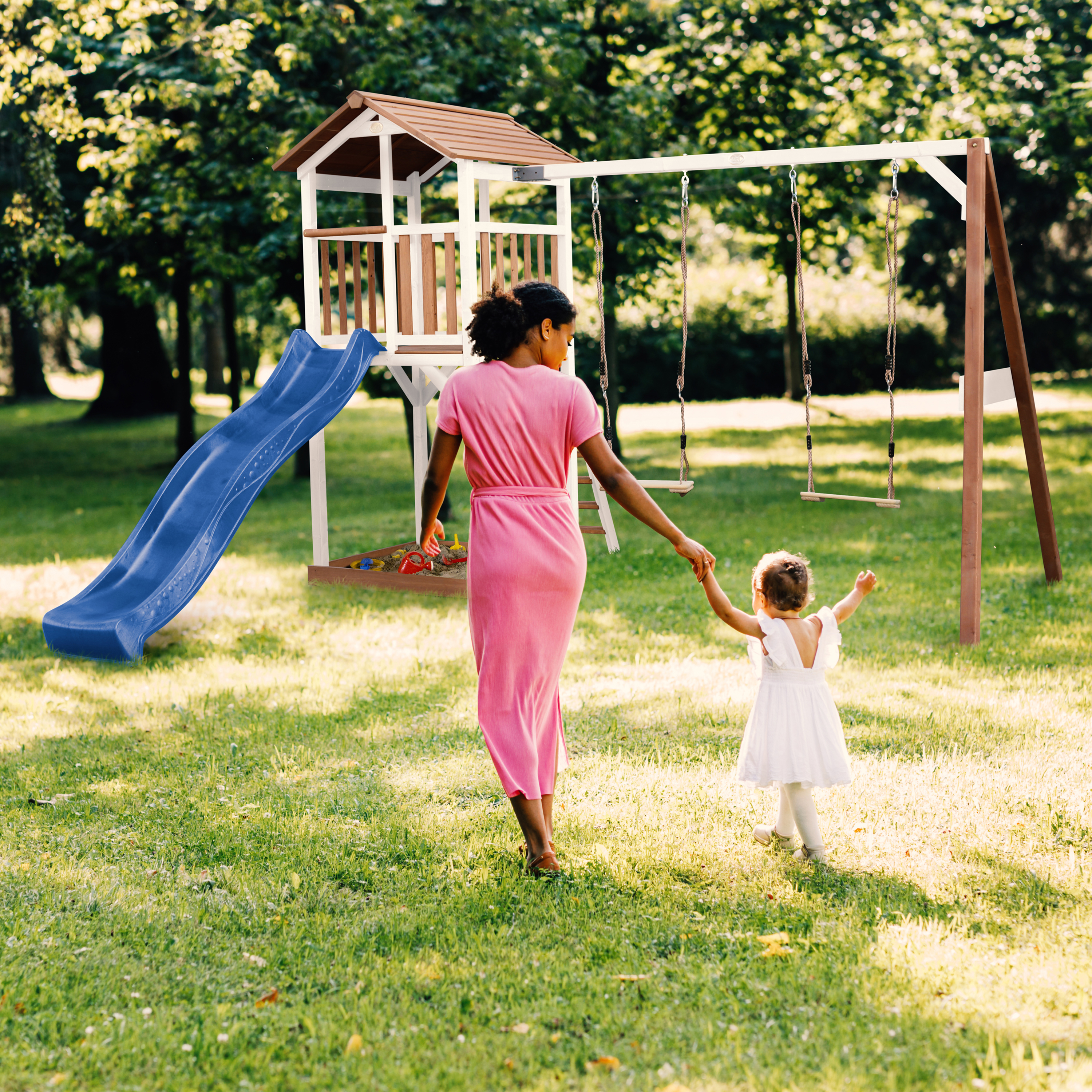 Mutter und Kind auf einem Spielplatz mit Rutsche und Schaukeln im Gras.