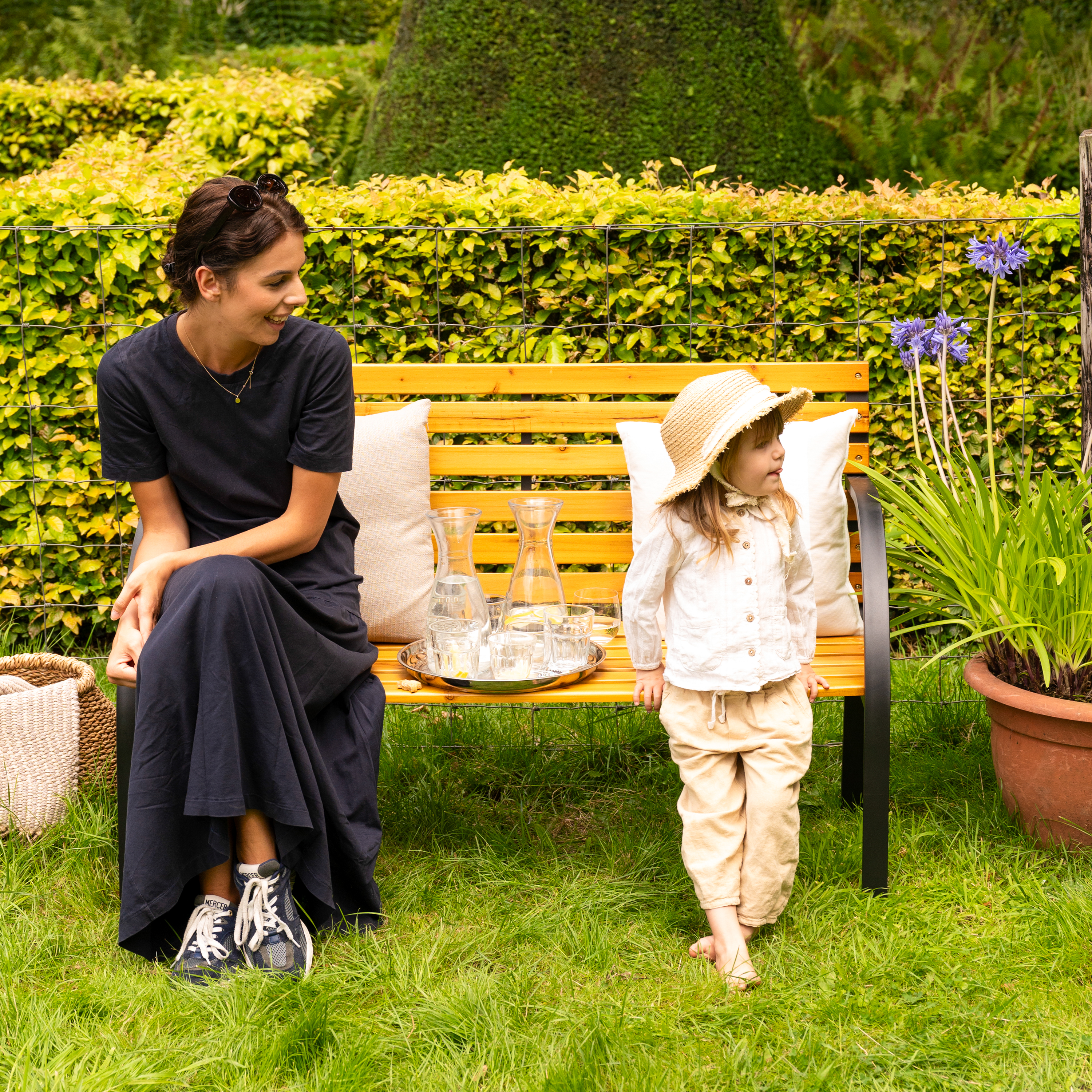 Frau und Kind sitzen auf einer Bank im Garten mit Getränken und Blumen.