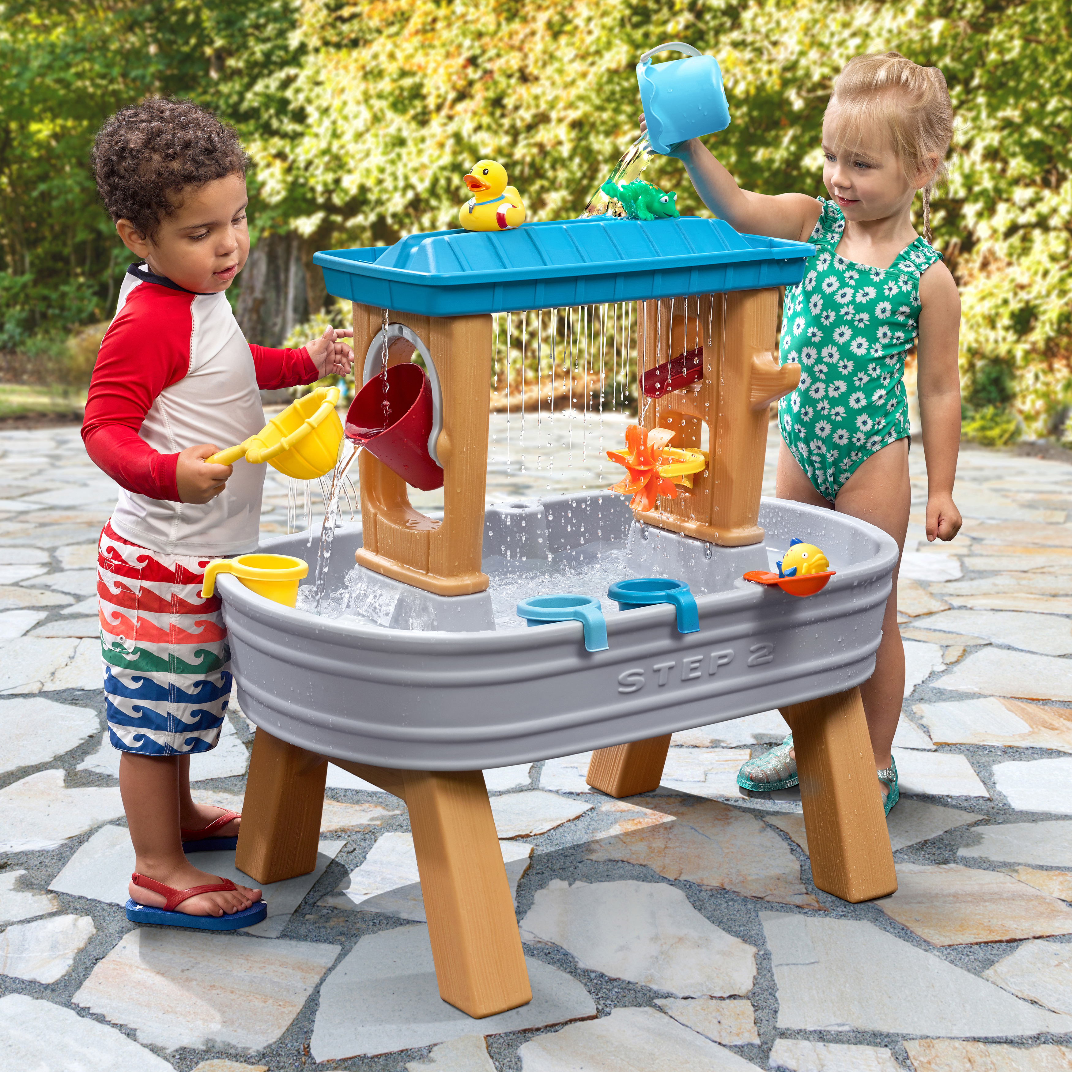 Zwei Kinder spielen mit einem Wasserspieltisch im Freien. Ein Junge gießt Wasser ein, ein Mädchen gießt Wasser aus einem blauen Eimer.
