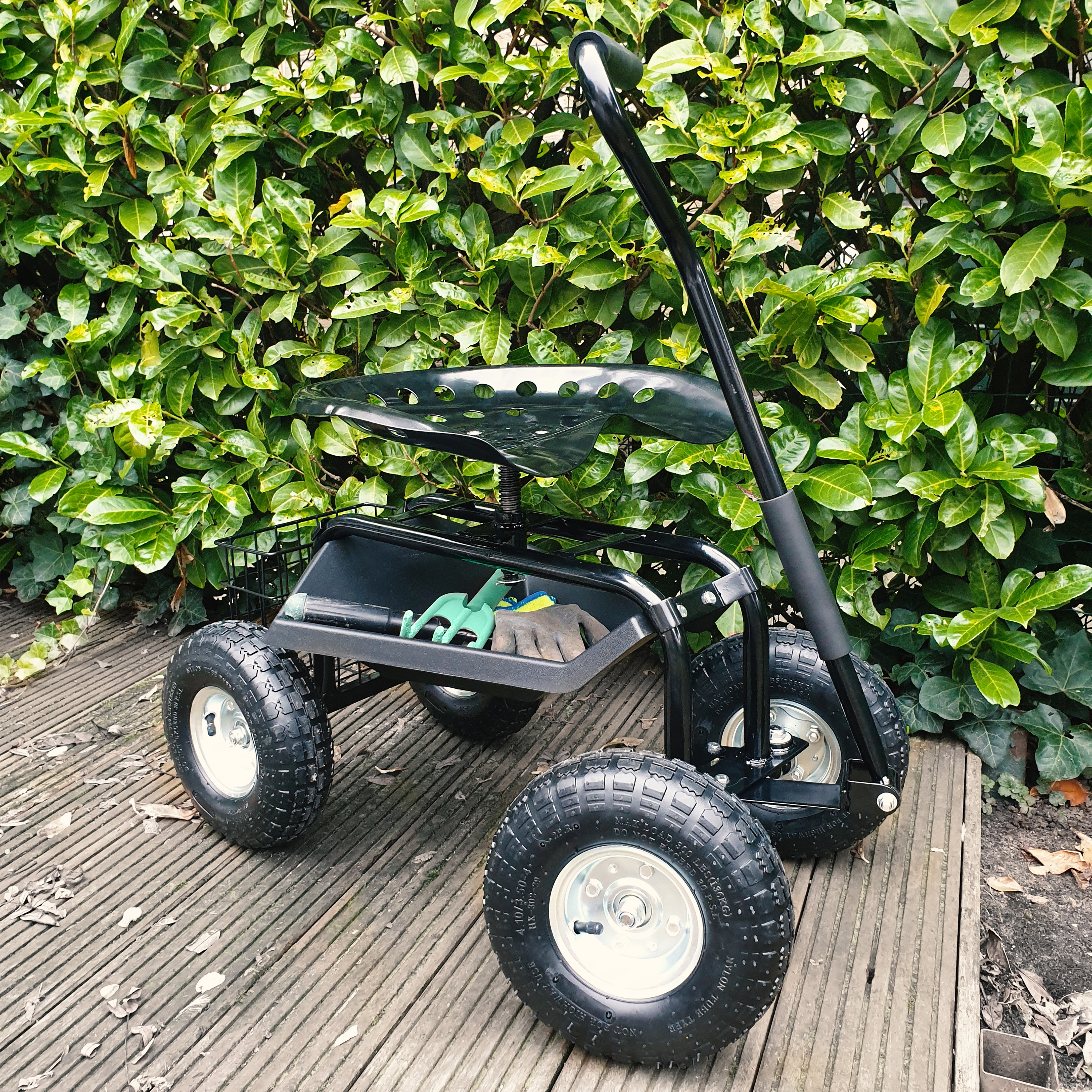 Ein schwarzer Gartensitzroller mit vier Rädern steht auf einem Holzdeck vor einer grünen Hecke.