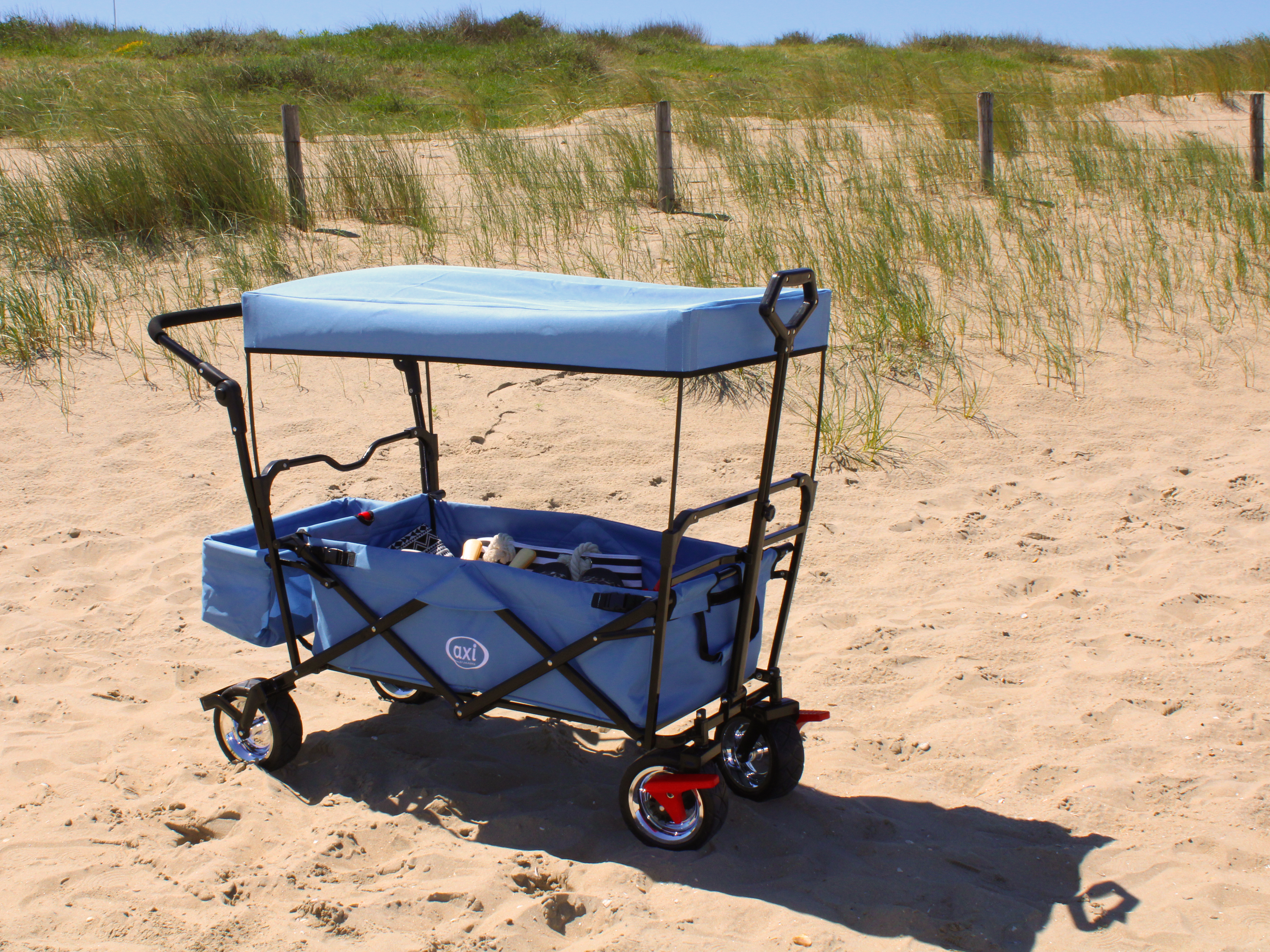Ein blauer Strandwagen mit Dach steht auf sandigem Boden. Im Hintergrund Gras und Holzpfähle.