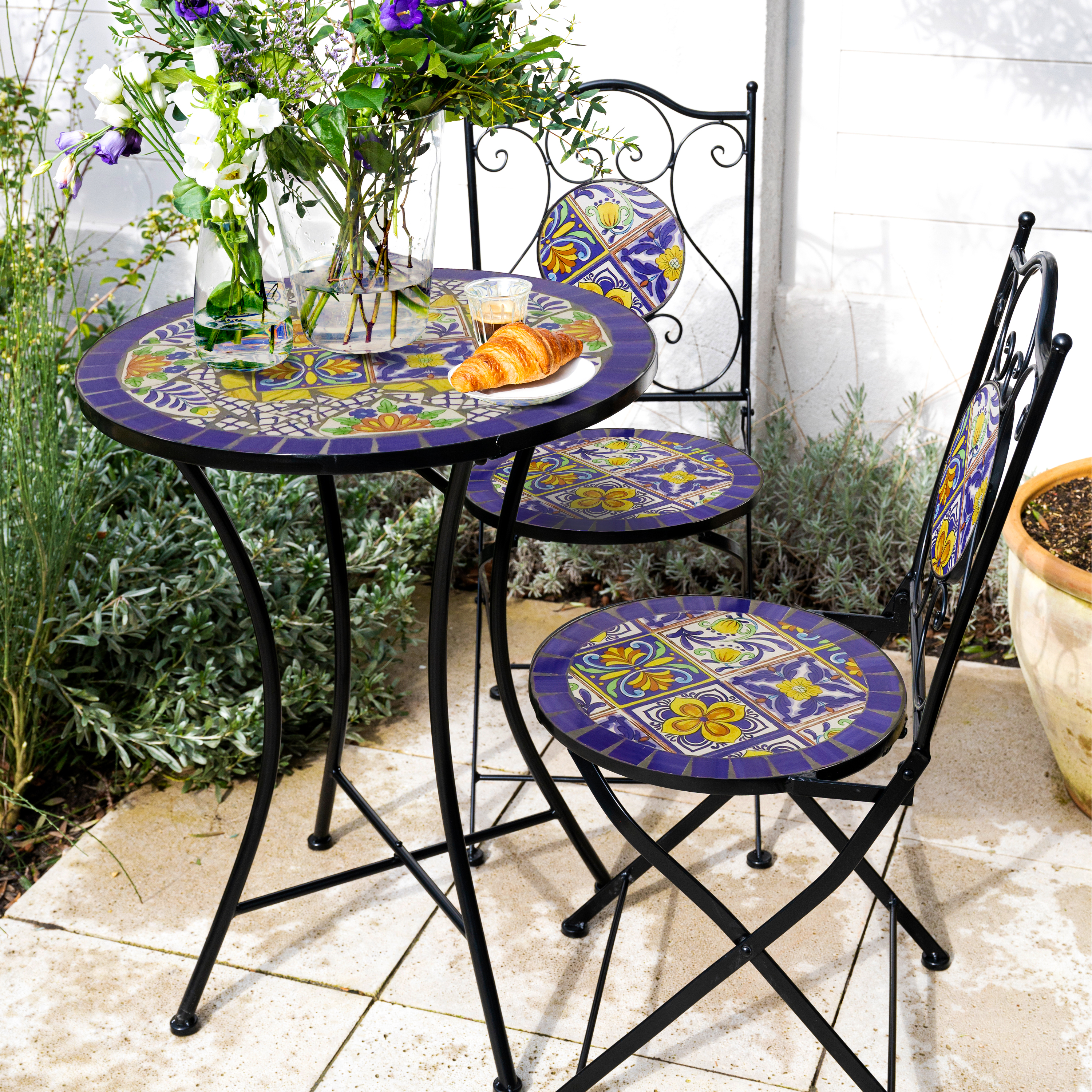 Mosaiktisch und Stühle im Freien mit Blumen und einem Croissant.