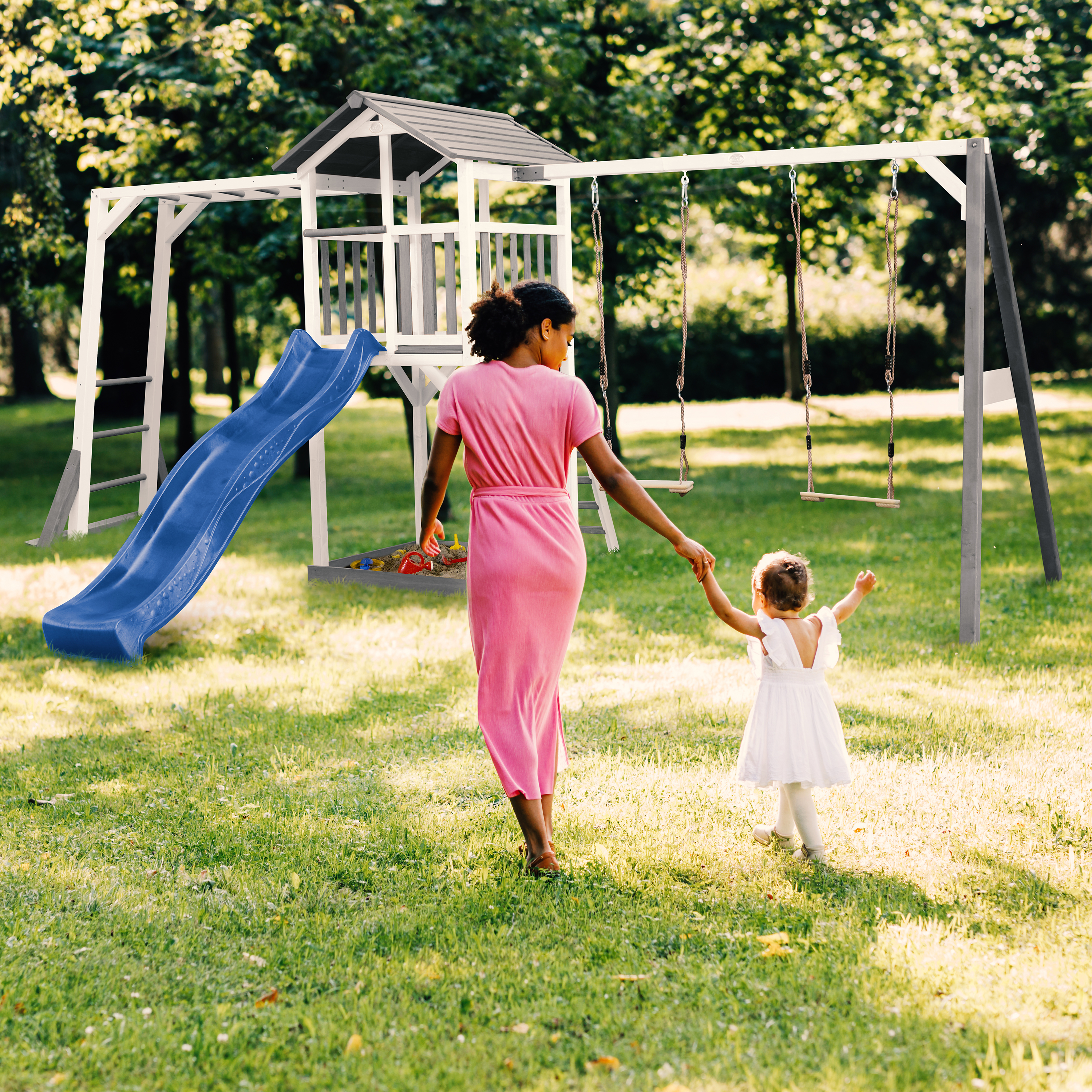 Mutter und Kind gehen zu einem Spielturm mit Rutsche und Schaukeln im Garten.