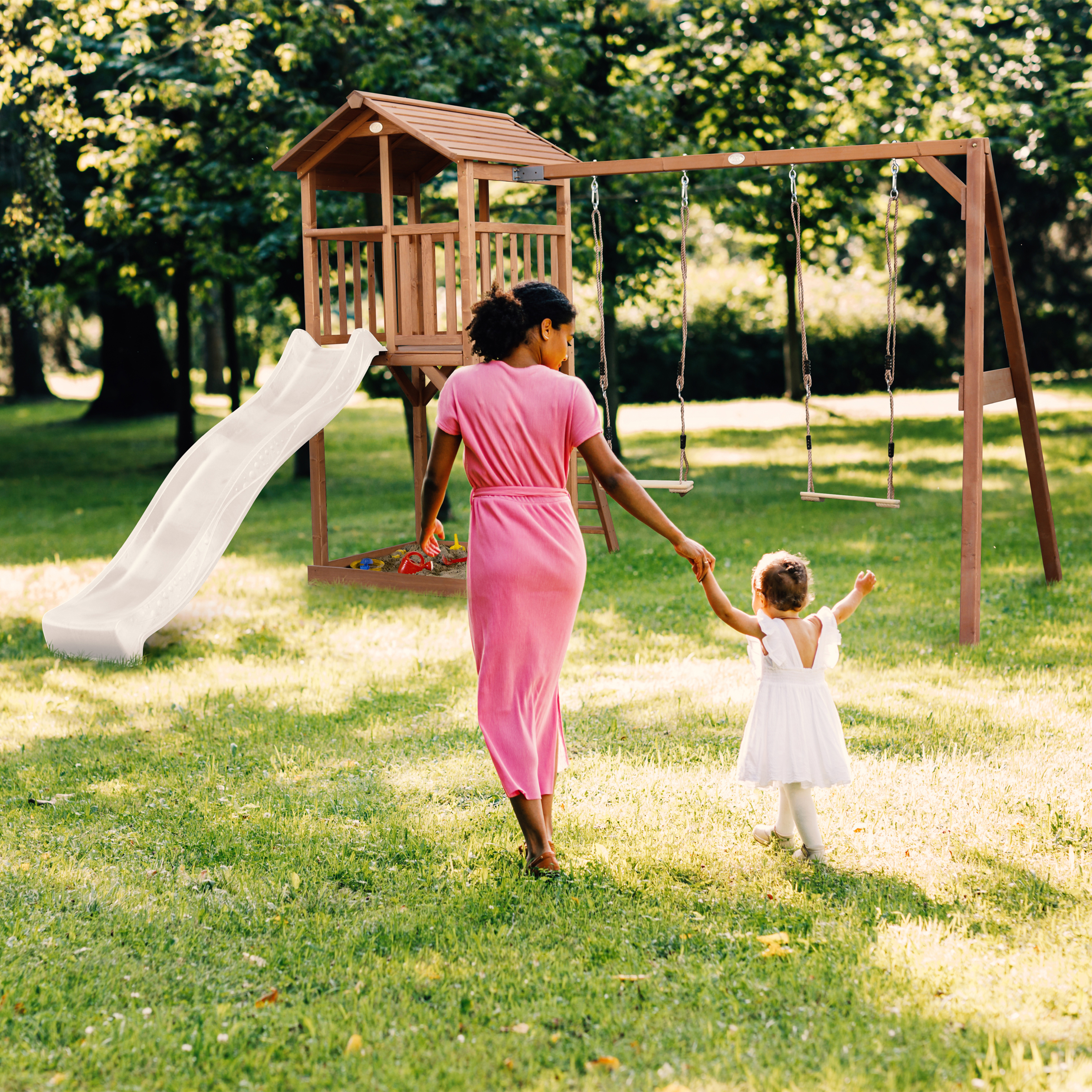 Mutter und Kind gehen zu einem Spielturm mit Rutsche und Schaukeln im Garten.