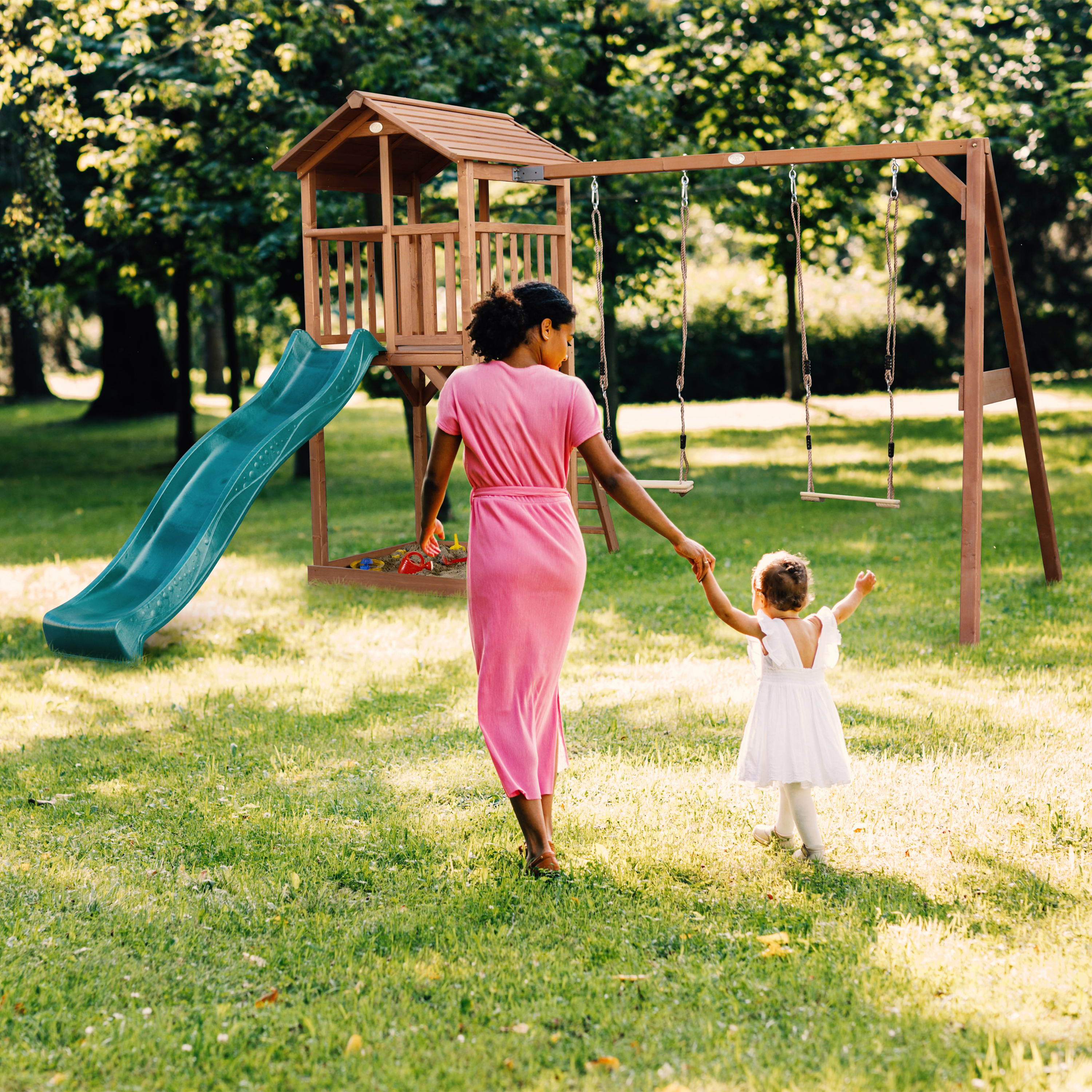 Frau mit Kind im Garten, Spielturm mit Rutsche und Schaukeln im Hintergrund.