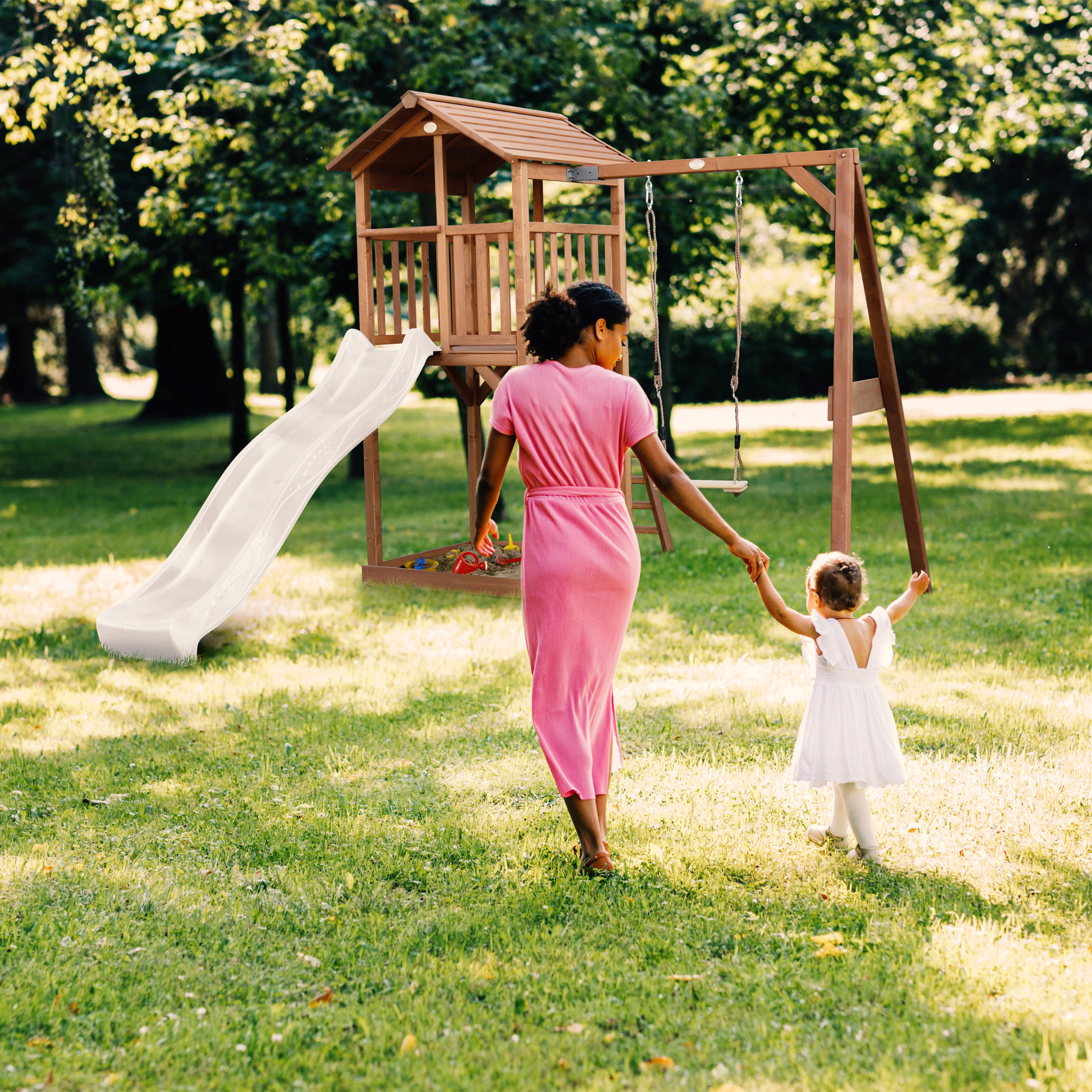 Frau in rosa Kleid und Kind in weißem Kleid gehen zum Spielturm mit Rutsche und Schaukel.