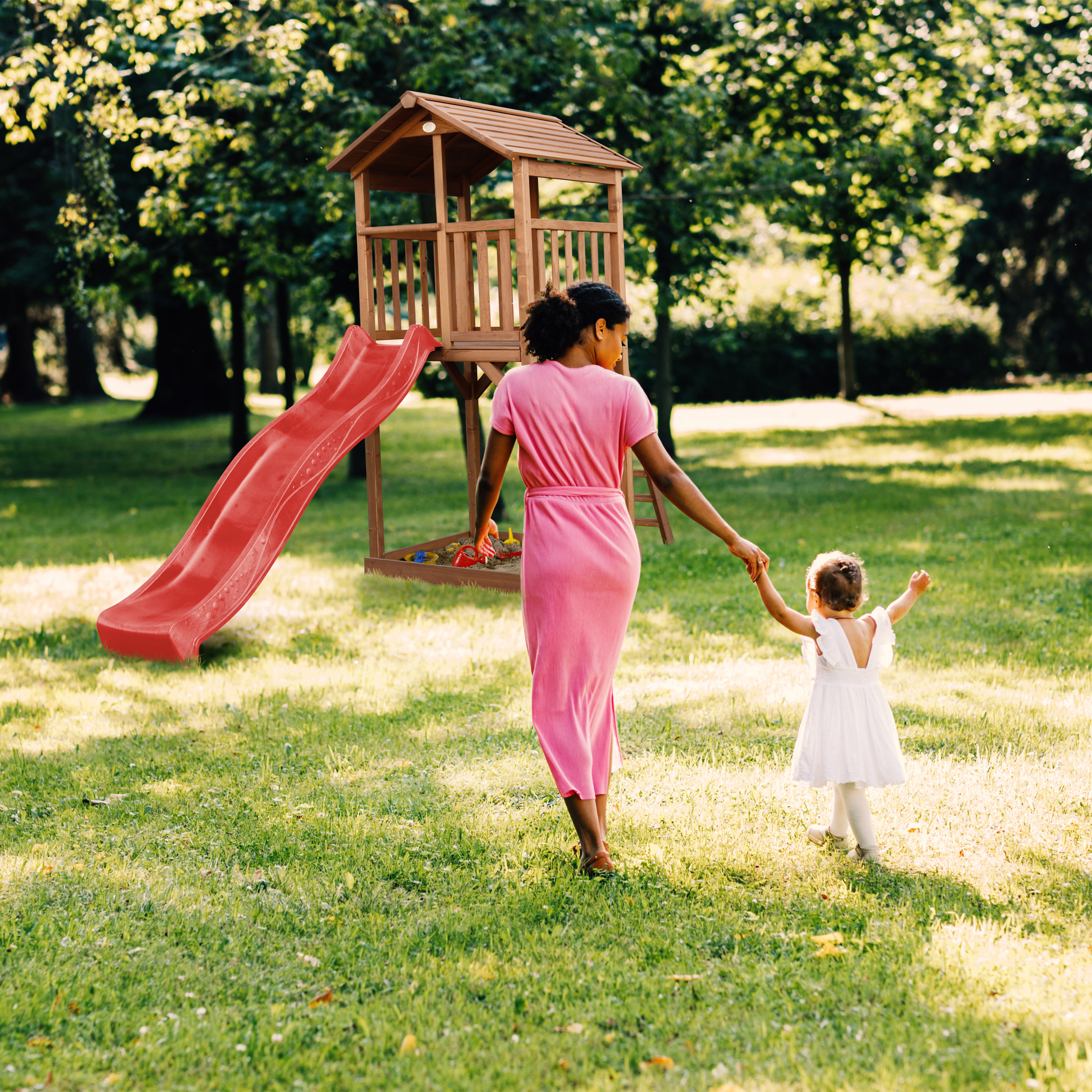 Eine Frau im rosa Kleid führt ein Kind im weißen Kleid zu einem Spielturm mit Rutsche.