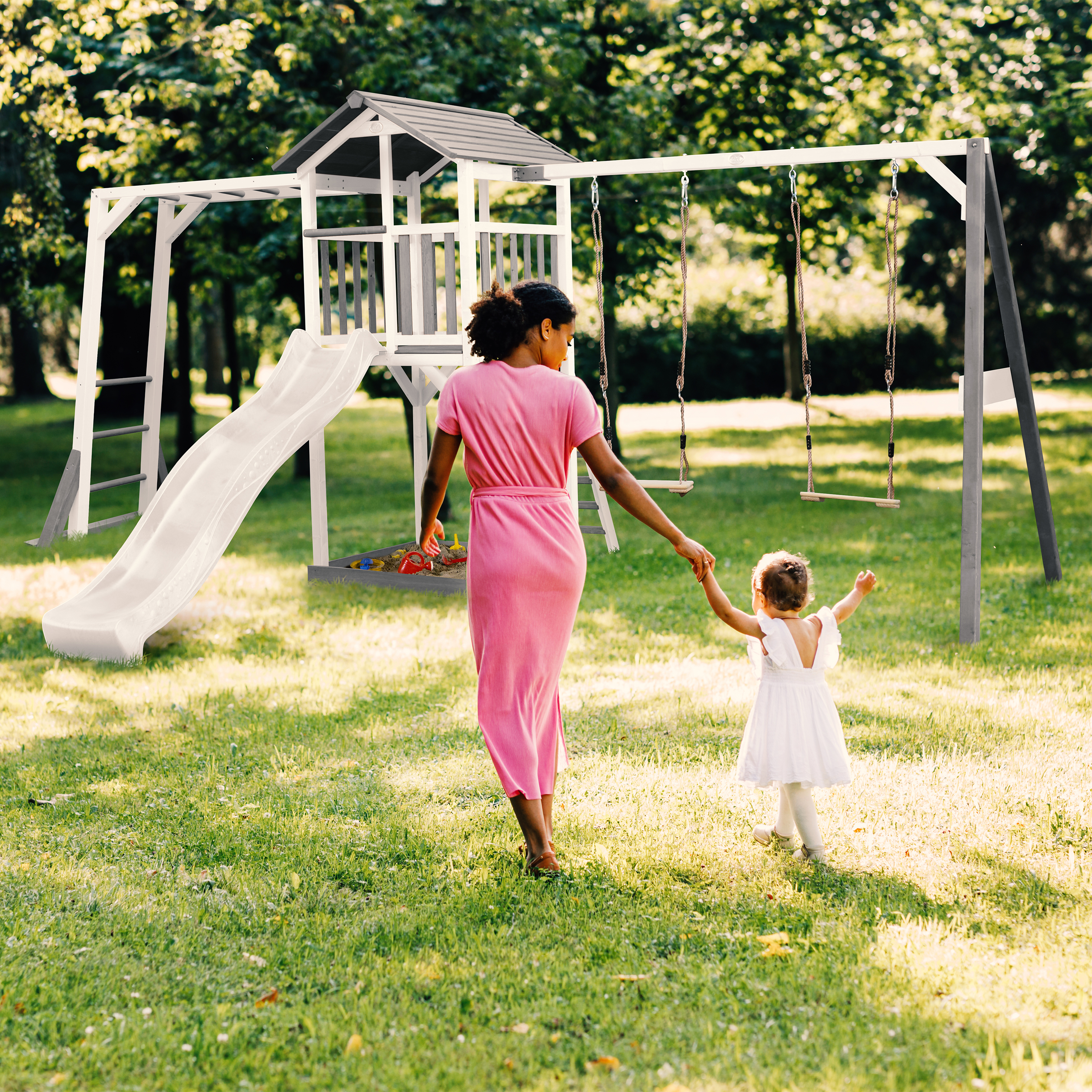 Mutter und Kind gehen auf Spielplatz mit Rutsche und Schaukeln im Gras.