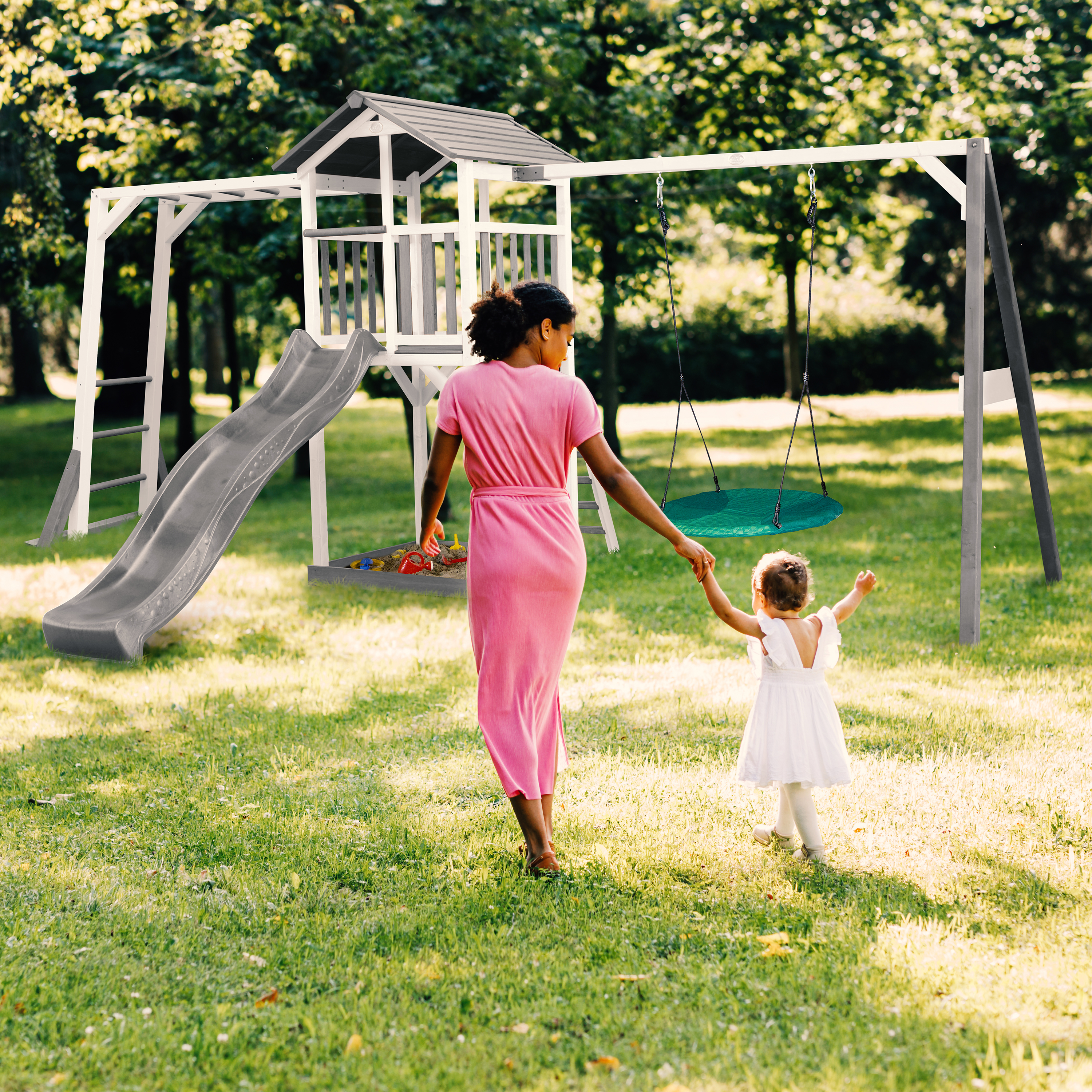 Frau mit Kind geht auf Spielplatz mit Rutsche und Schaukel im Gras.