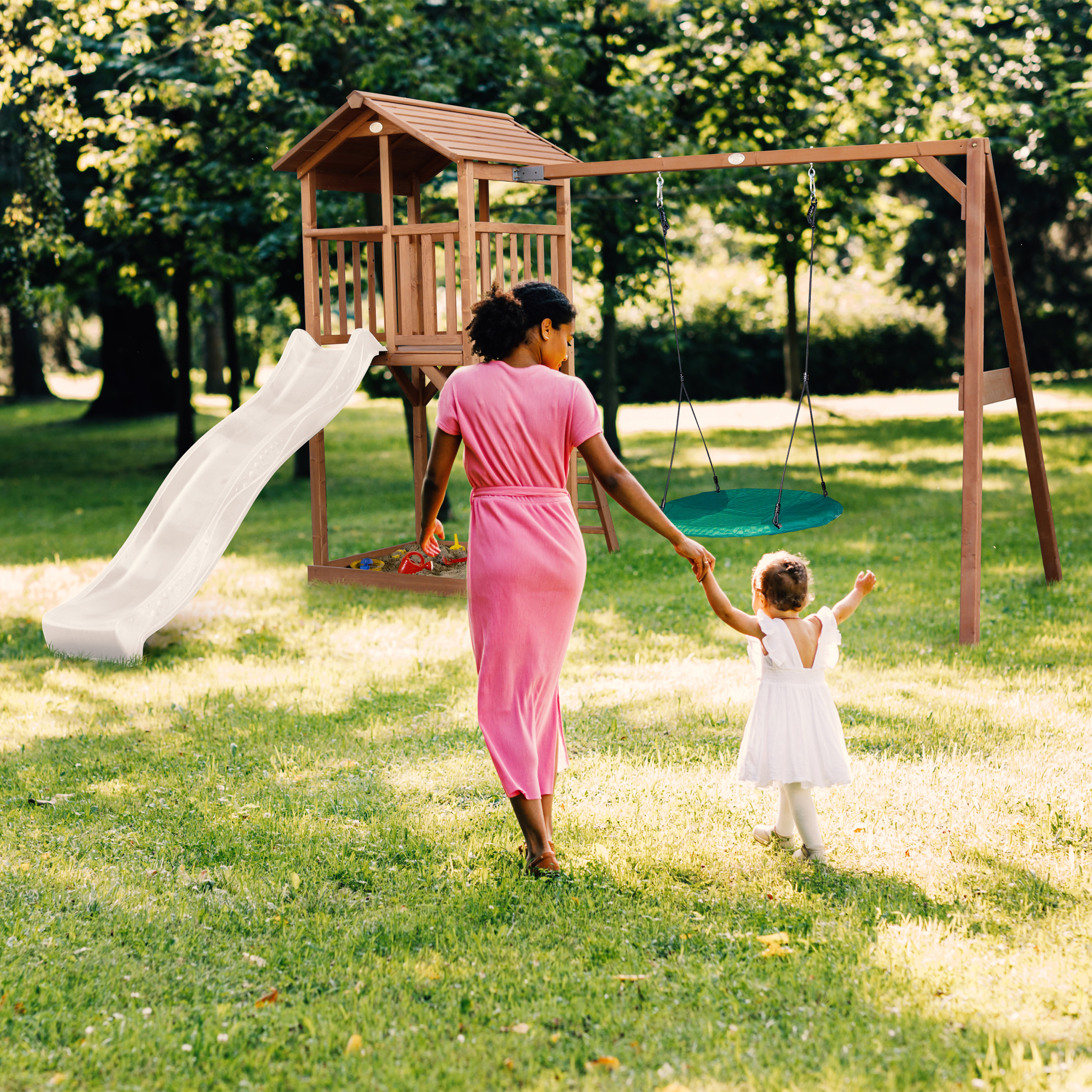 Eine Frau in einem rosa Kleid und ein Kind in Weiß gehen auf einen Holzspielplatz zu.