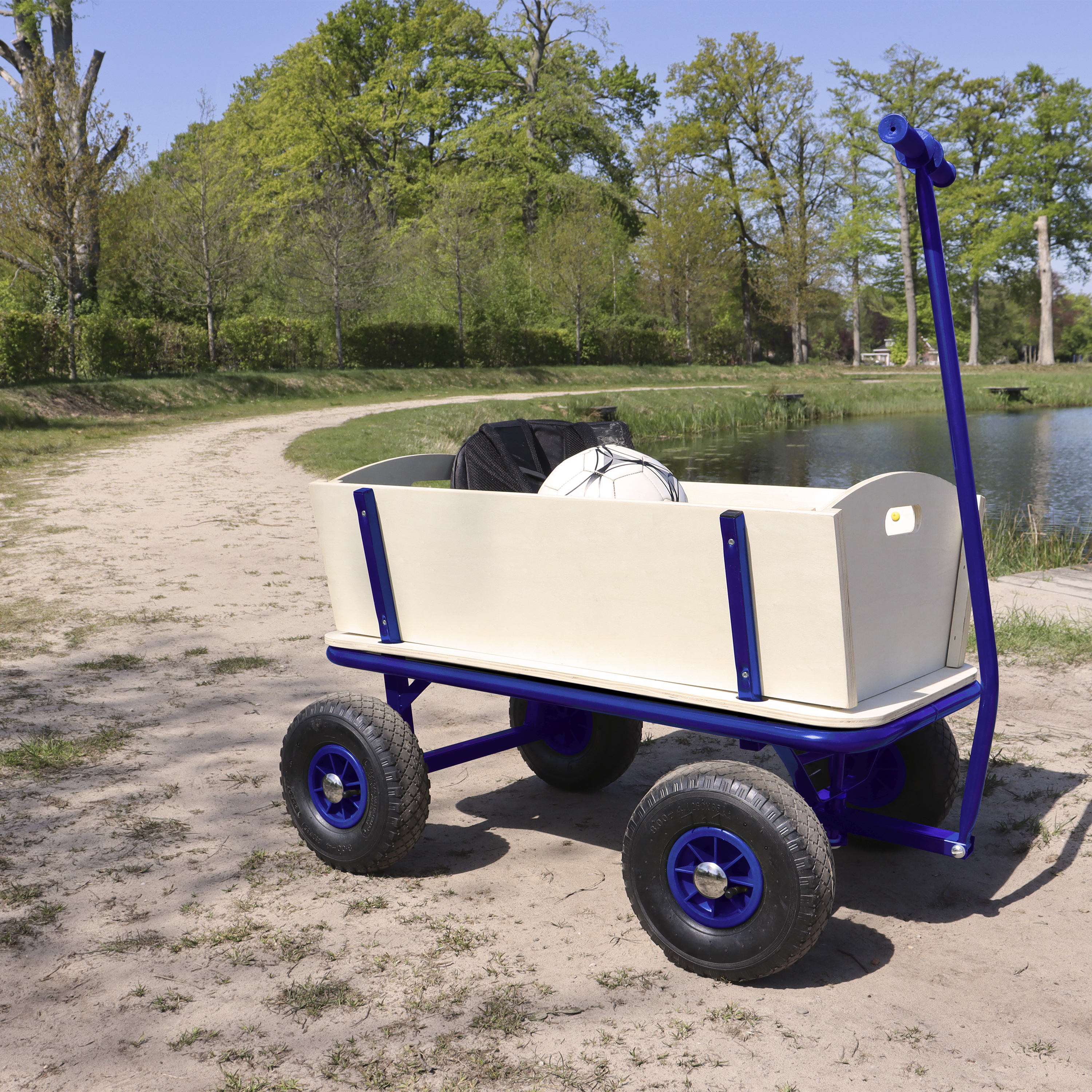 Ein blauer Bollerwagen mit Holzaufbau steht auf Sand neben einem See.