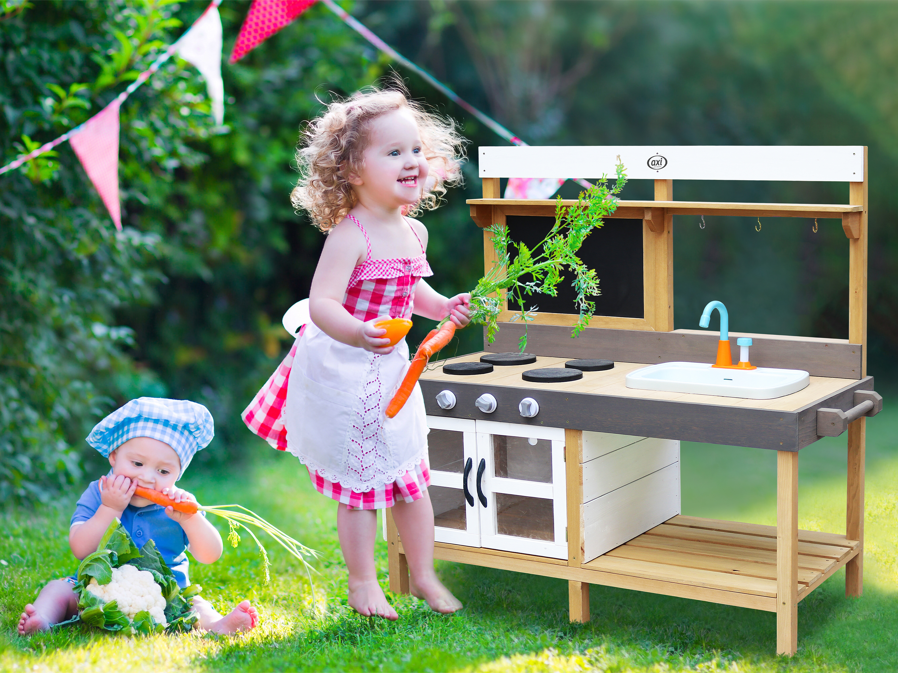 Zwei Kinder spielen im Garten mit einer Spielküche und Gemüse.