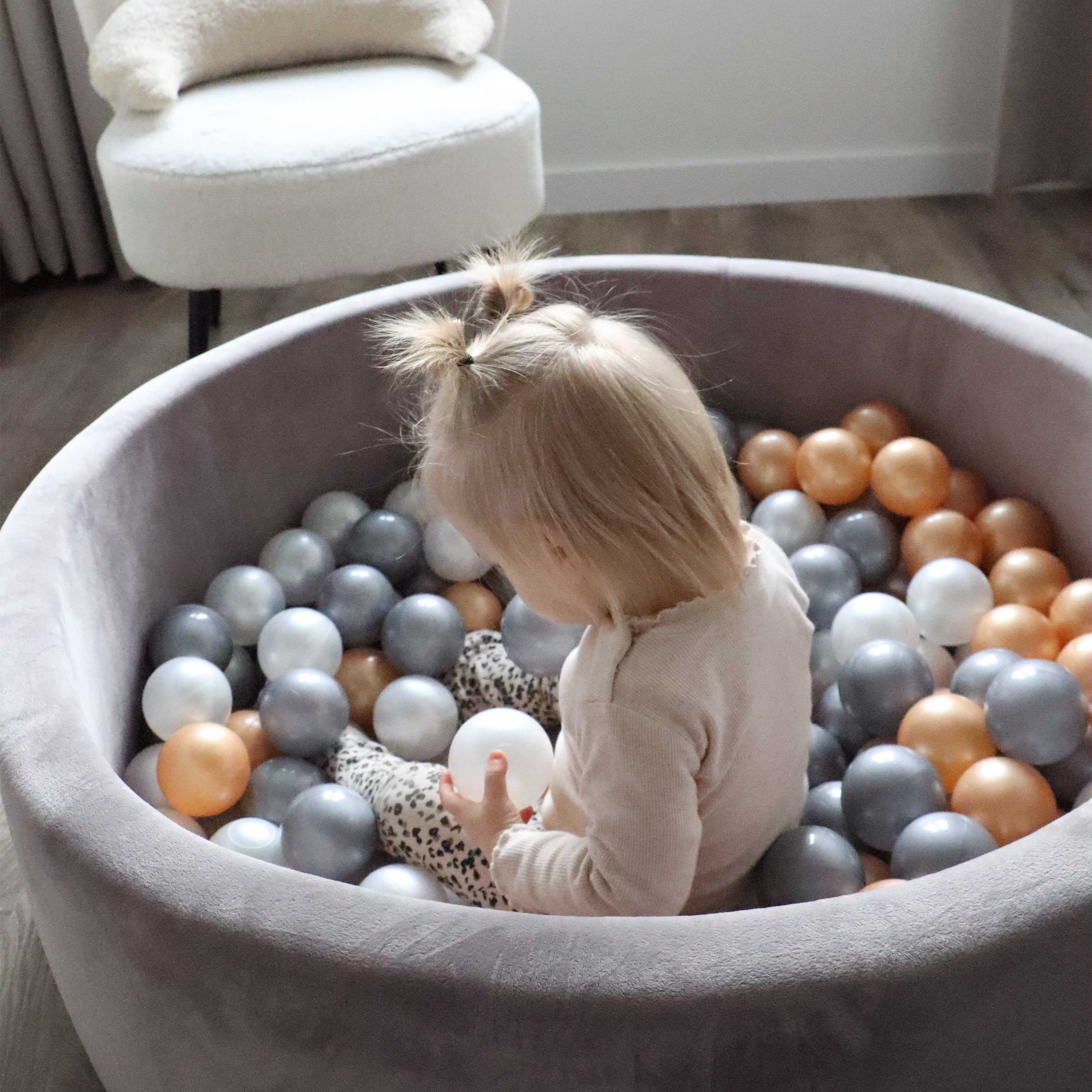 Ein Kleinkind sitzt in einem Ballbad mit grauen, weißen und orangen Bällen.