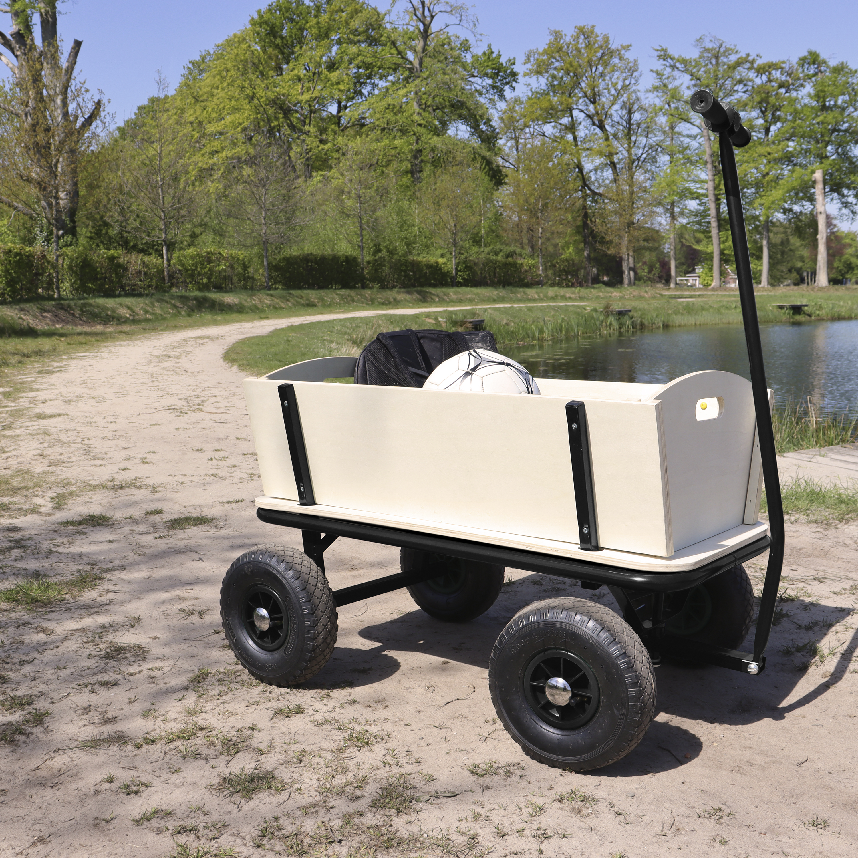 Ein heller Bollerwagen mit schwarzen Rädern steht auf Sand, mit einem See und Bäumen im Hintergrund.