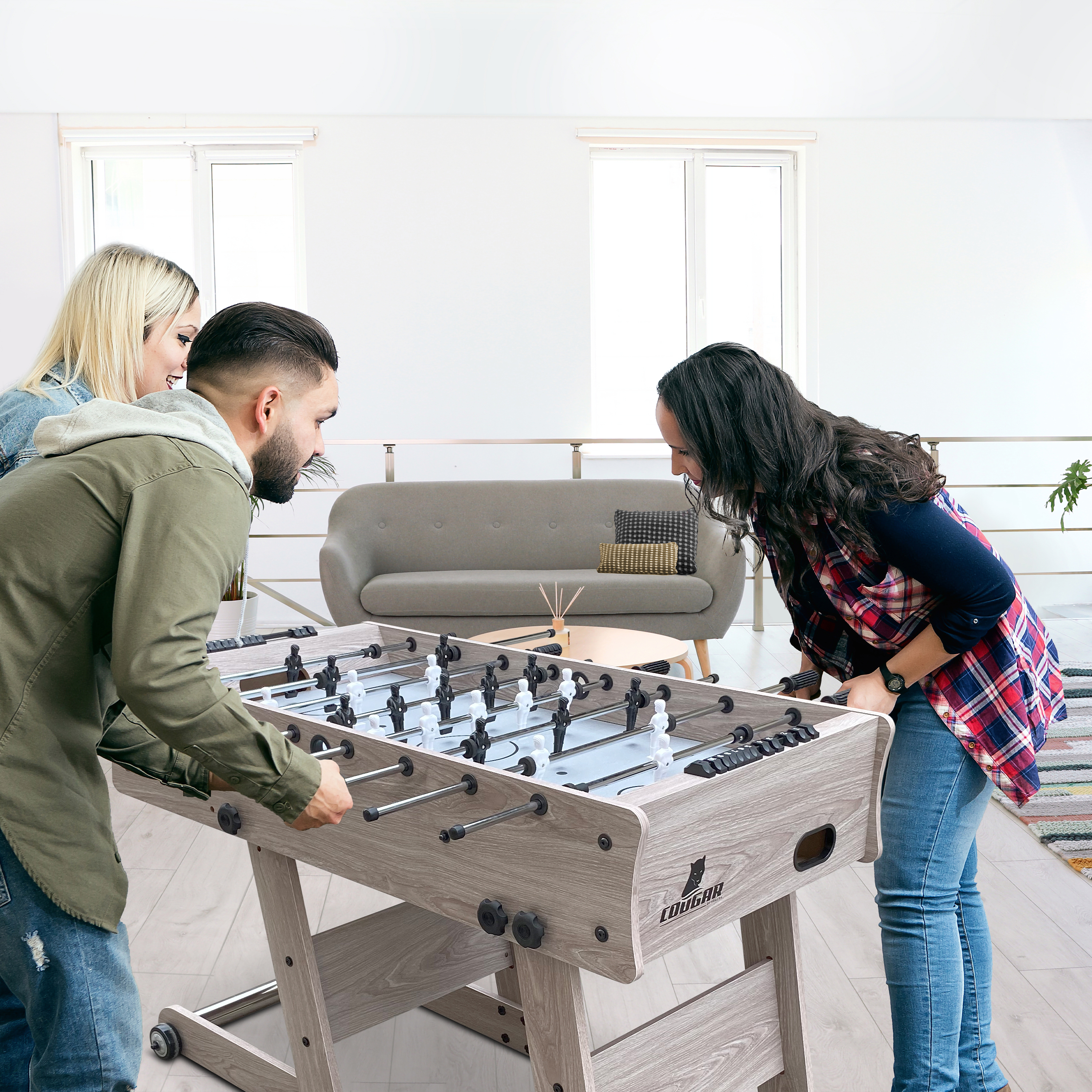 Drei Personen spielen Tischfußball in einem hellen Raum mit einem Sofa im Hintergrund.