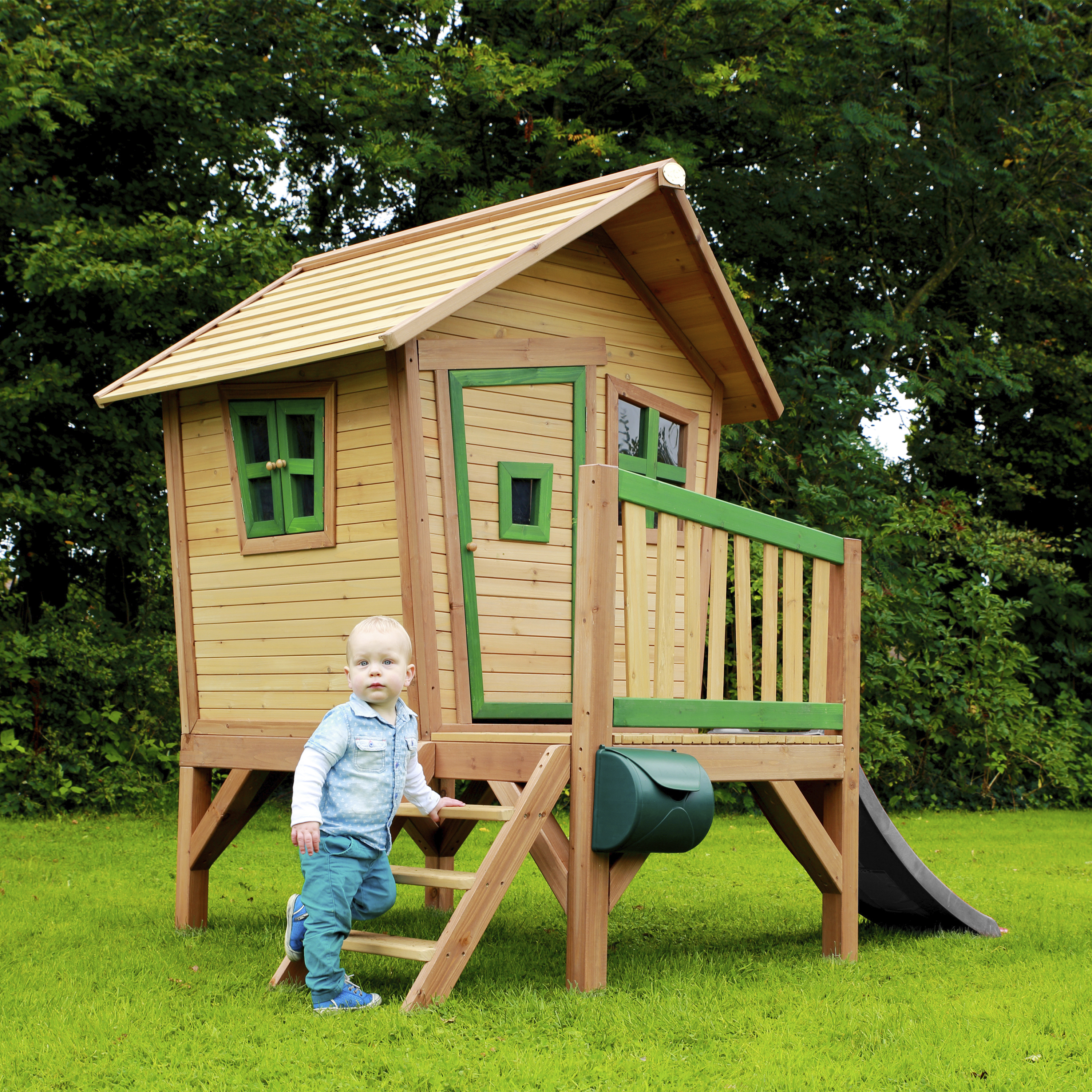 Ein Kleinkind steht auf den Stufen vor einem Holzspielhaus im Grünen.