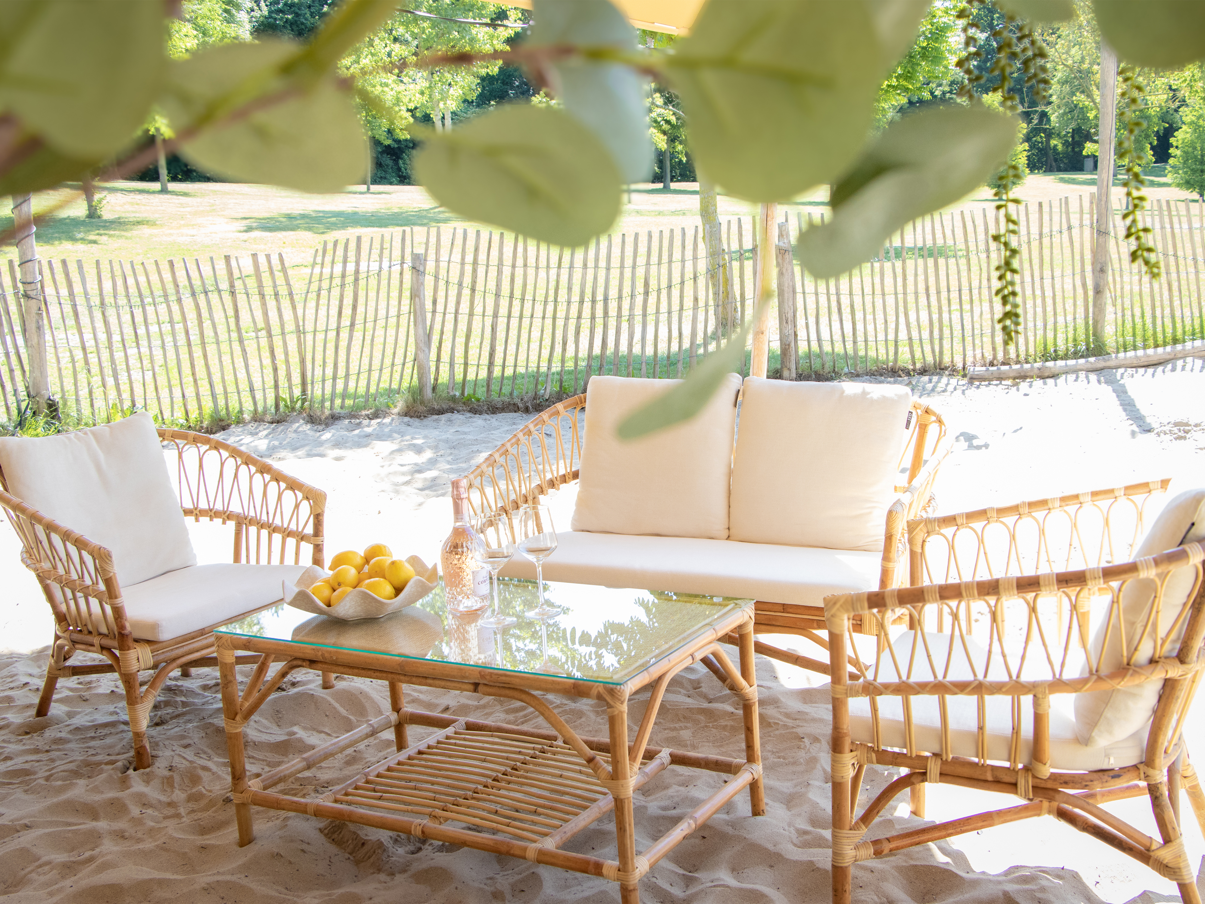 Rattan-Möbel mit weißen Kissen am Strand, daneben ein Tisch mit Zitronen und Wein.