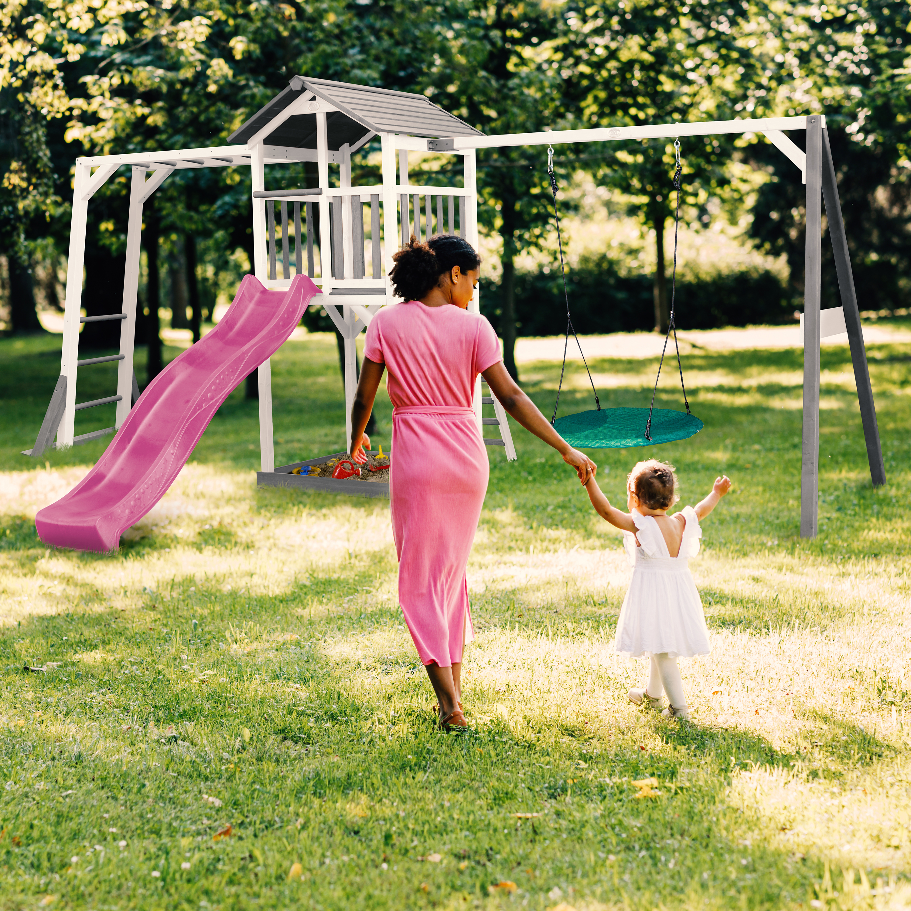 Mutter und Kind gehen zum Spielplatz mit Rutsche und Schaukel im sonnigen Garten.