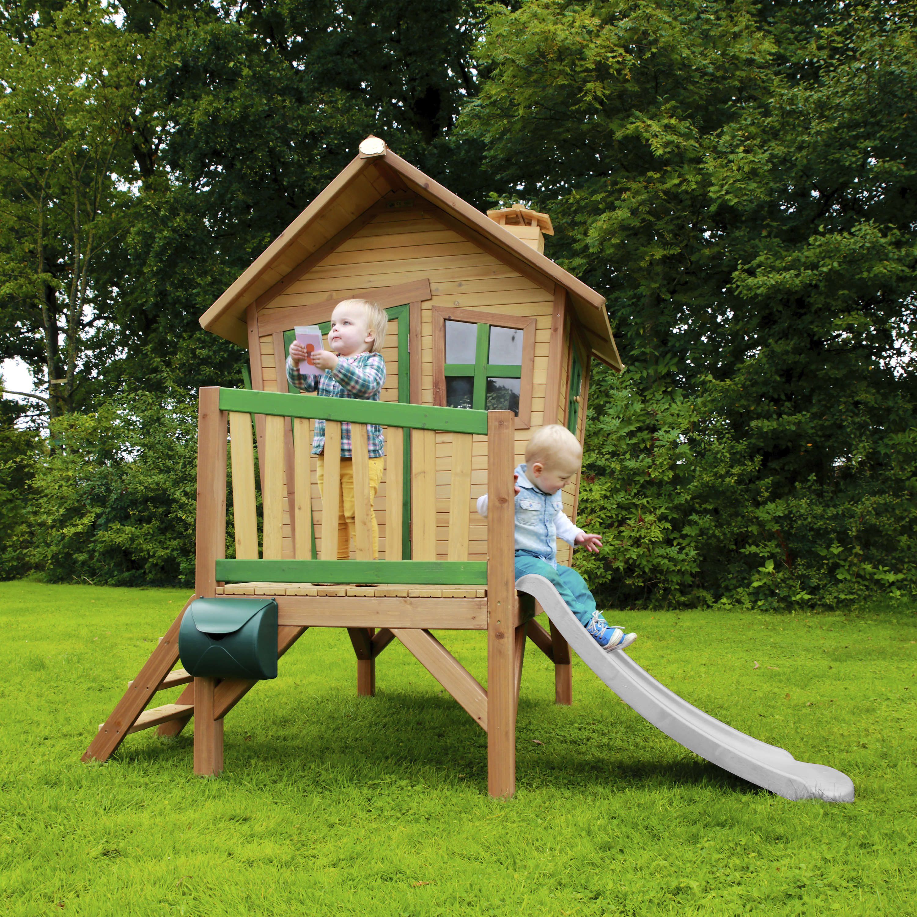 Zwei Kinder spielen auf einem Holzspielhaus mit Rutsche im Garten.