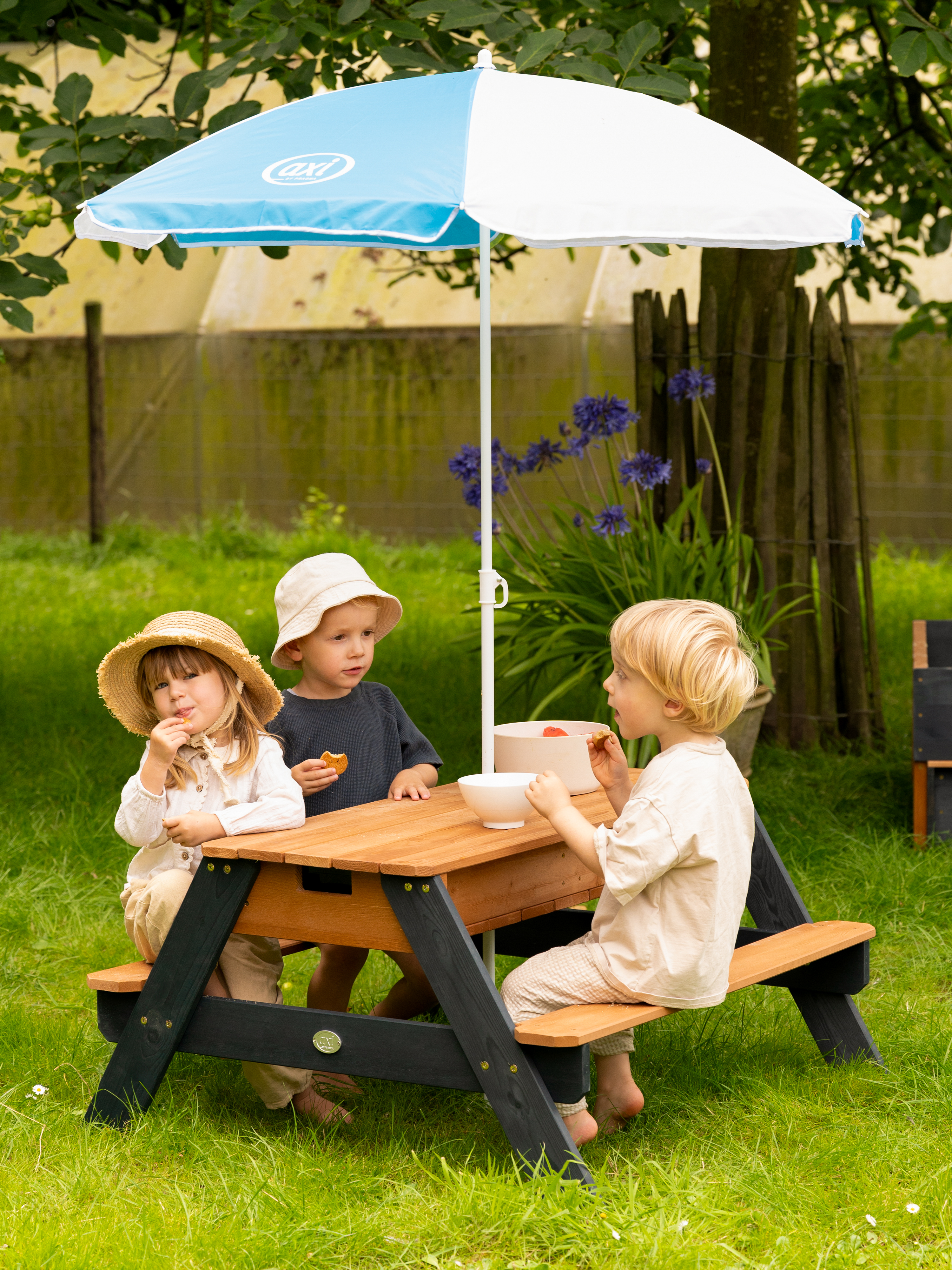 Drei Kinder sitzen an einem Holztisch im Garten unter einem Sonnenschirm.