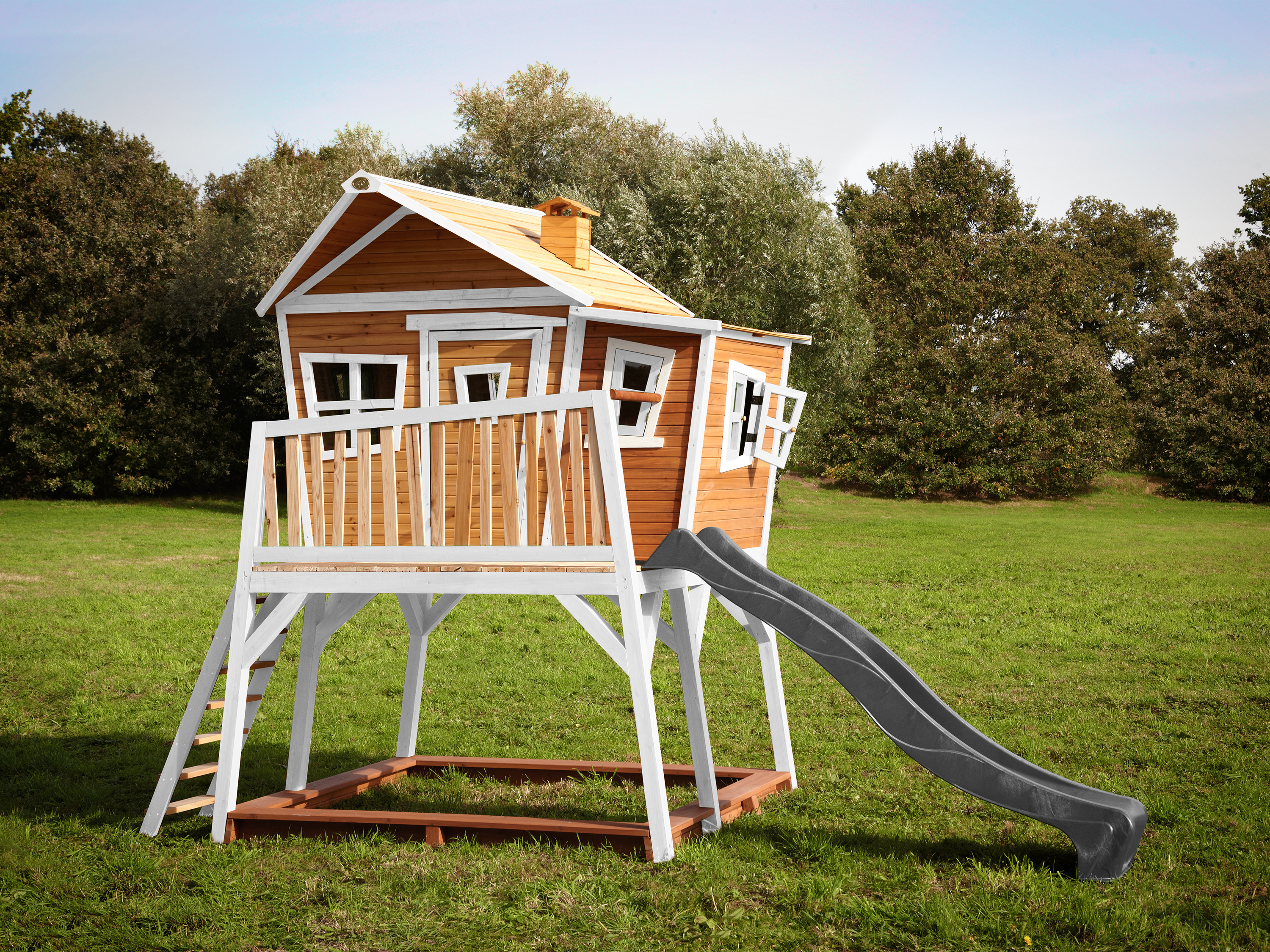 Ein hölzernes Spielhaus auf Stelzen mit Rutsche und Leiter auf grünem Gras.